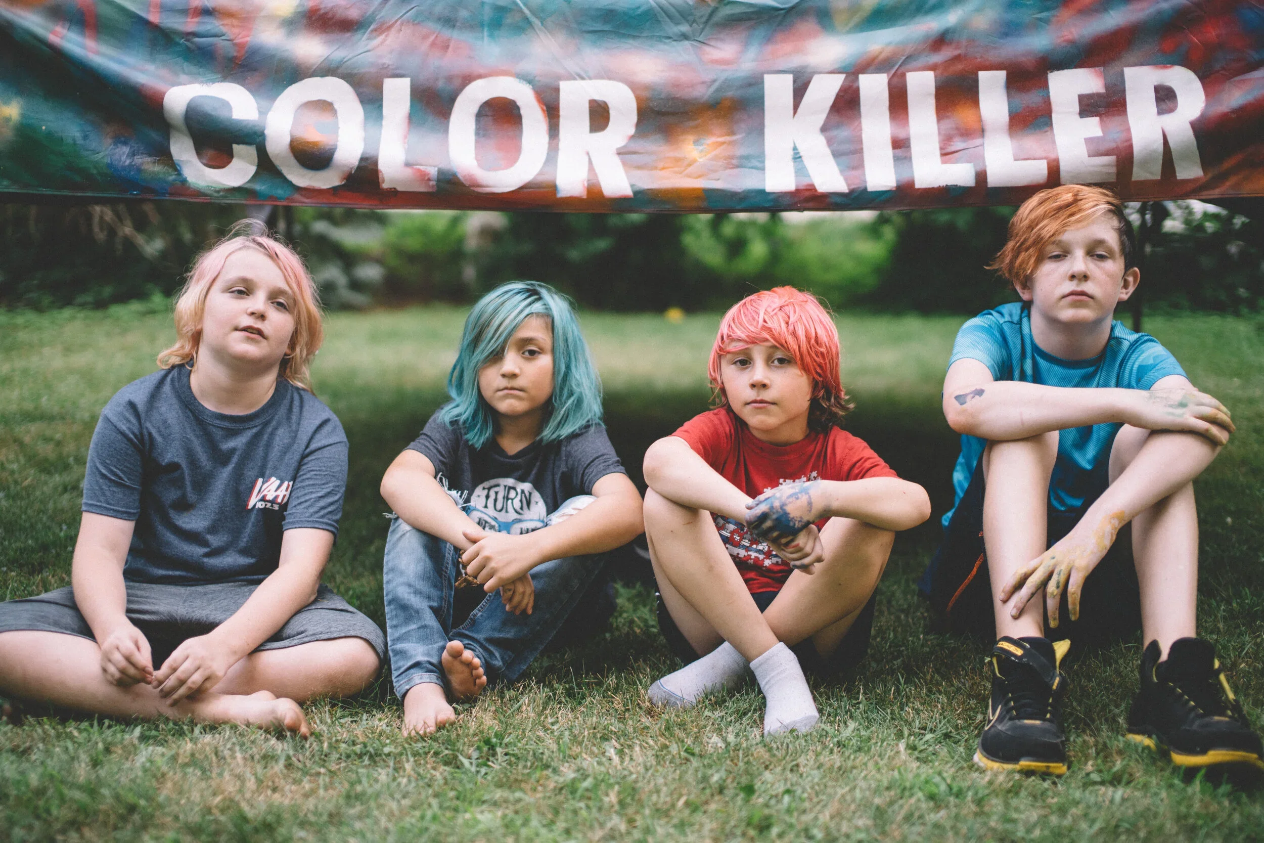 Four children sitting on grass under a colorful banner that reads 'COLOR KILLER', with paint on their hands and clothes.