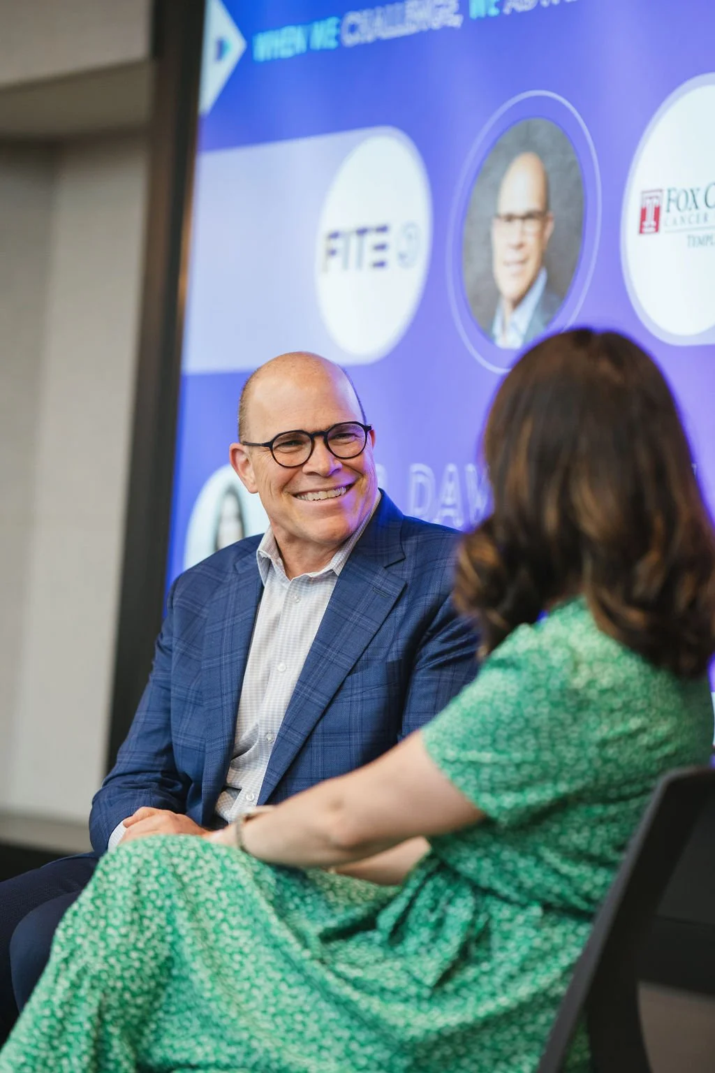 A man in a blue suit and glasses smiling and talking to a woman in a green dress during a panel discussion at a conference, with a screen displaying logos and a profile picture in the background.