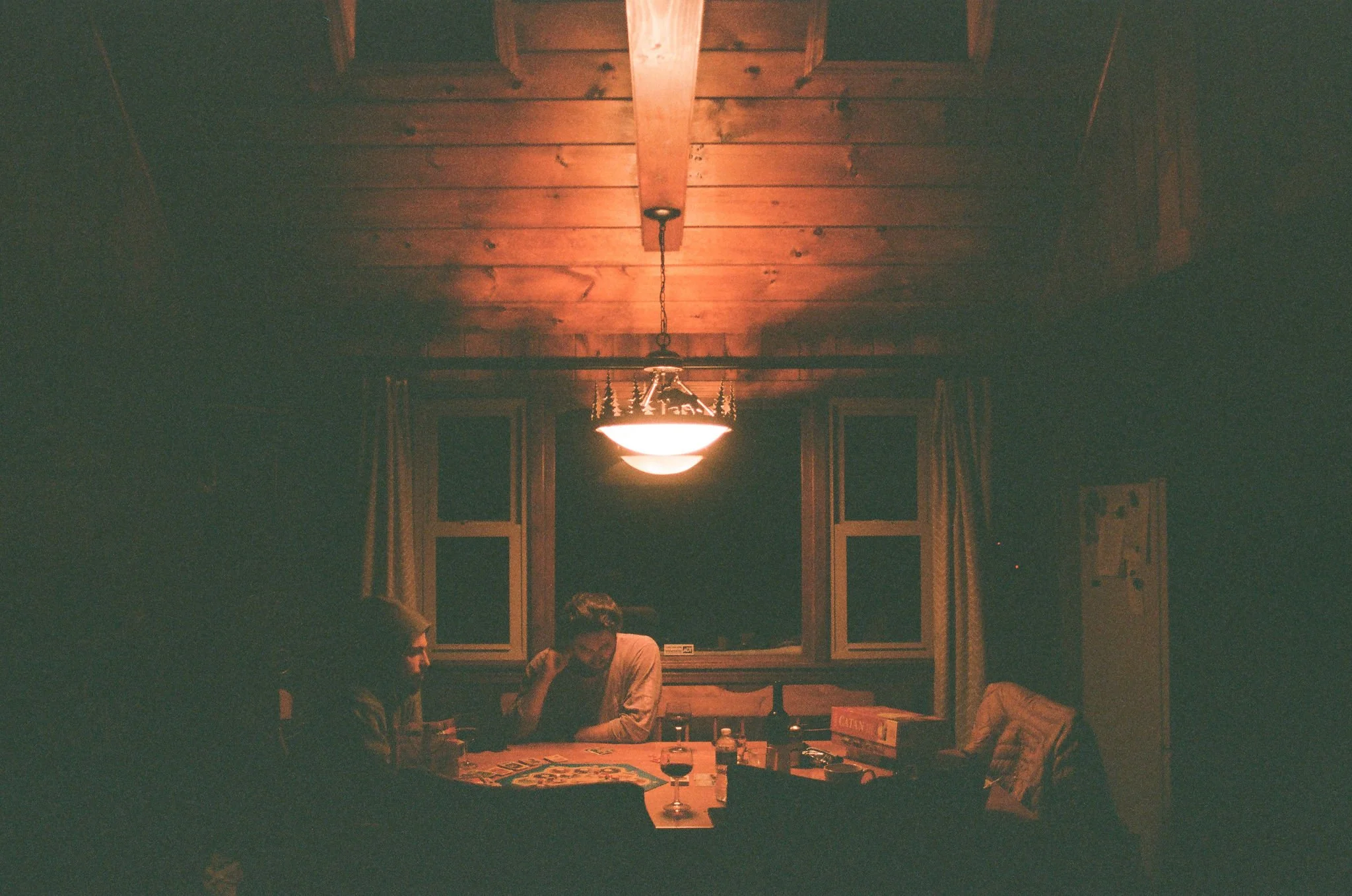A dimly lit wooden dining room with a hanging ceiling light, two people sitting at a table, and windows with curtains.