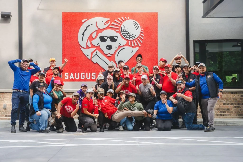 Group of people posing outdoors in front of a large red sign with a dog wearing sunglasses, a fish, a golf ball, and the word "SAUGUS".