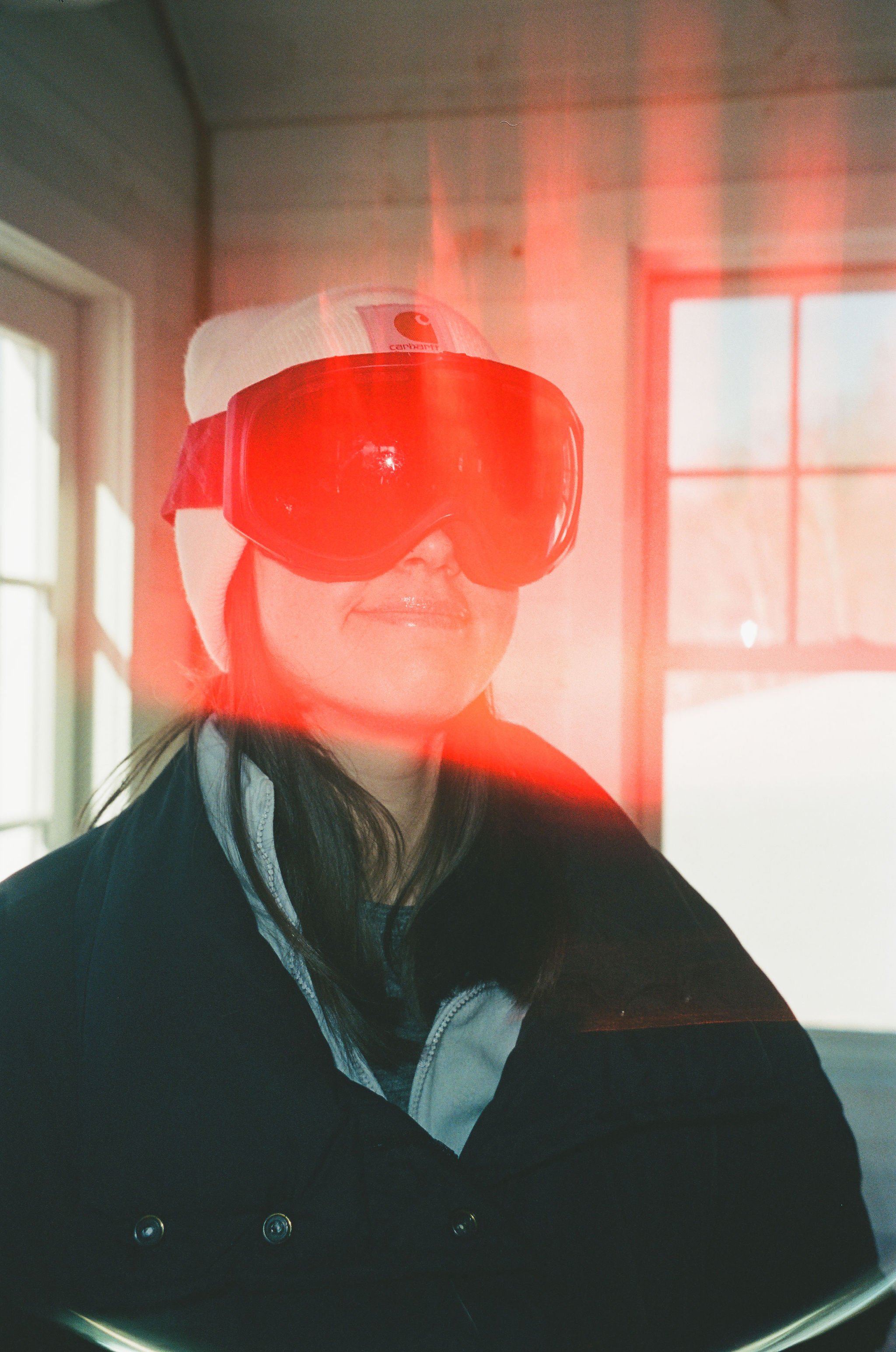A woman wearing a winter hat and ski goggles with a red glow reflection on the goggles, indoors near a window.