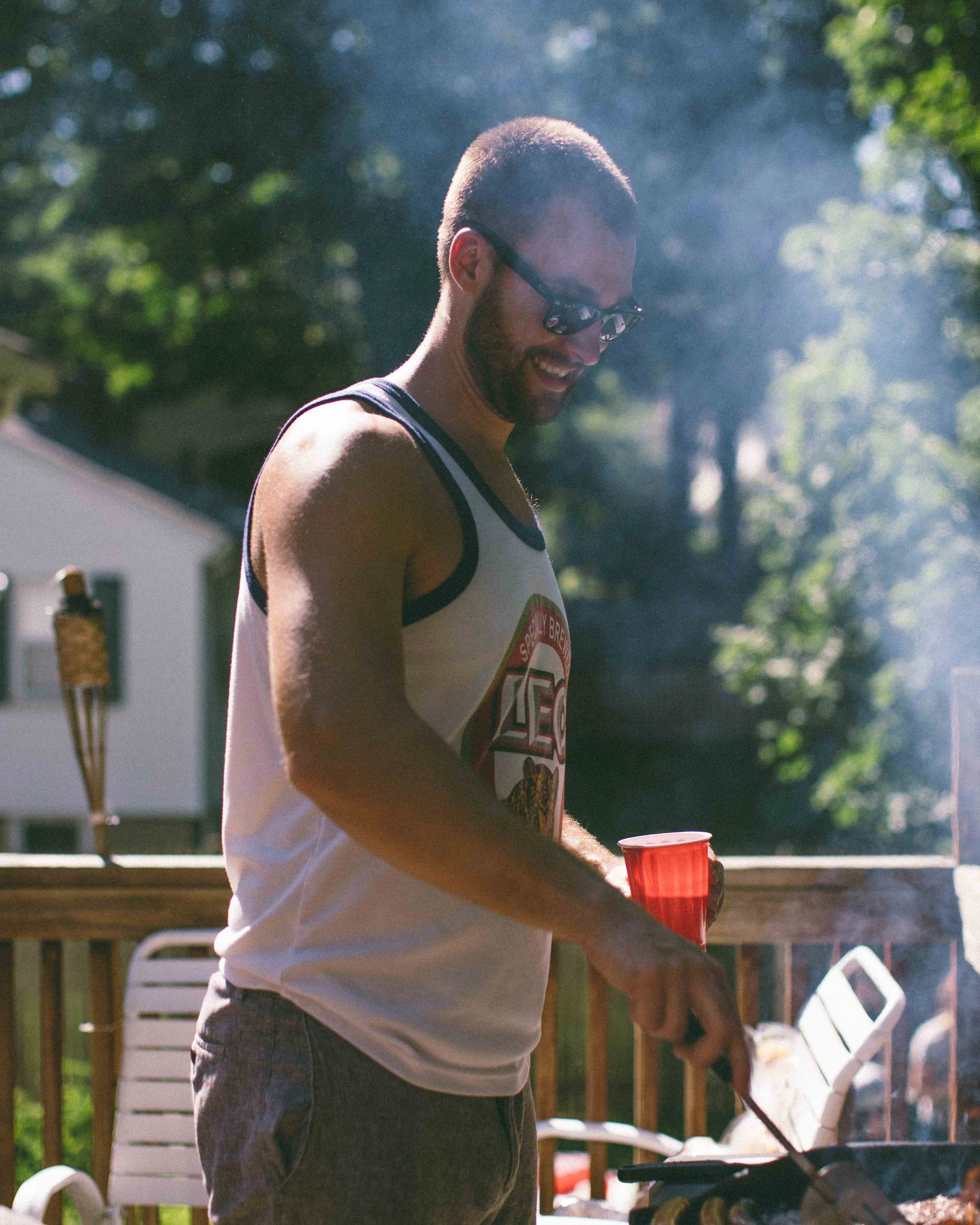 A man with glasses and a beard wearing a tank top grilling outdoors, holding a red cup, in a backyard with trees and sunlight.