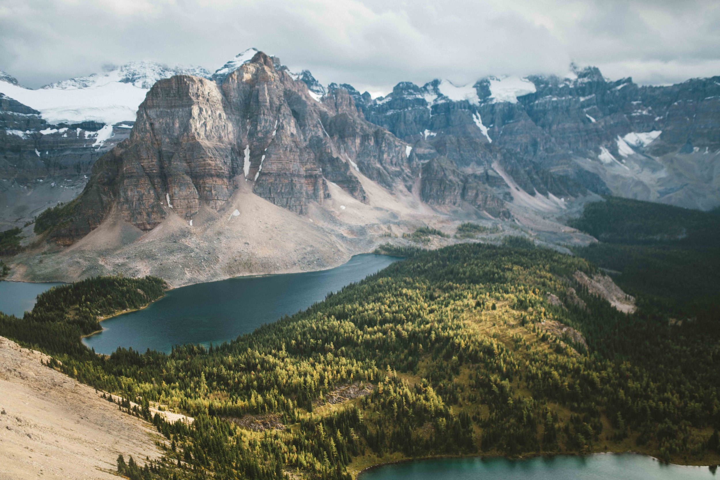 Snow-capped mountains with rocky peaks, a forested valley, and a lake in the foreground.