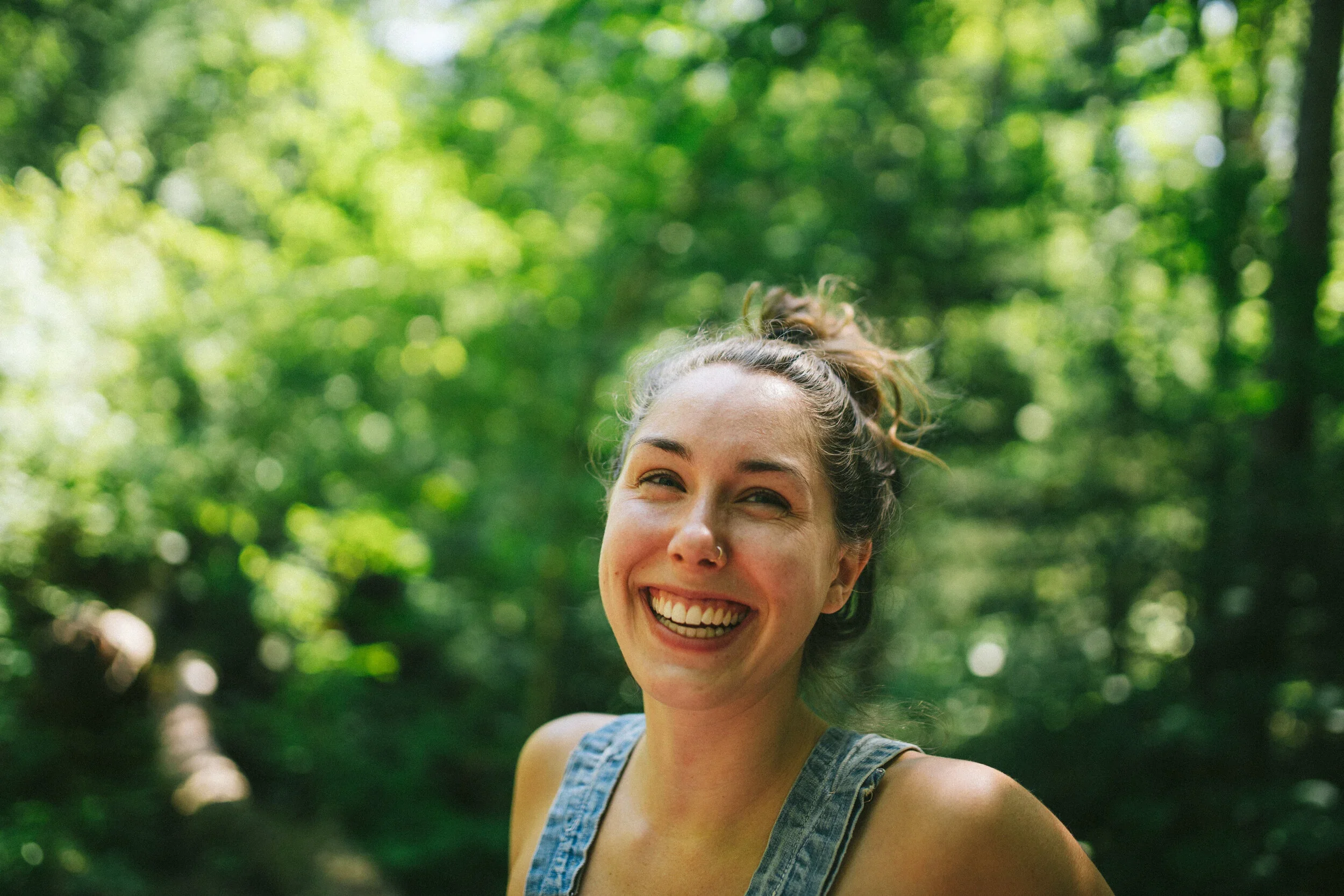 A young woman with a smile, wearing a tank top, standing outdoors in a green forest with sunlight filtering through the trees.