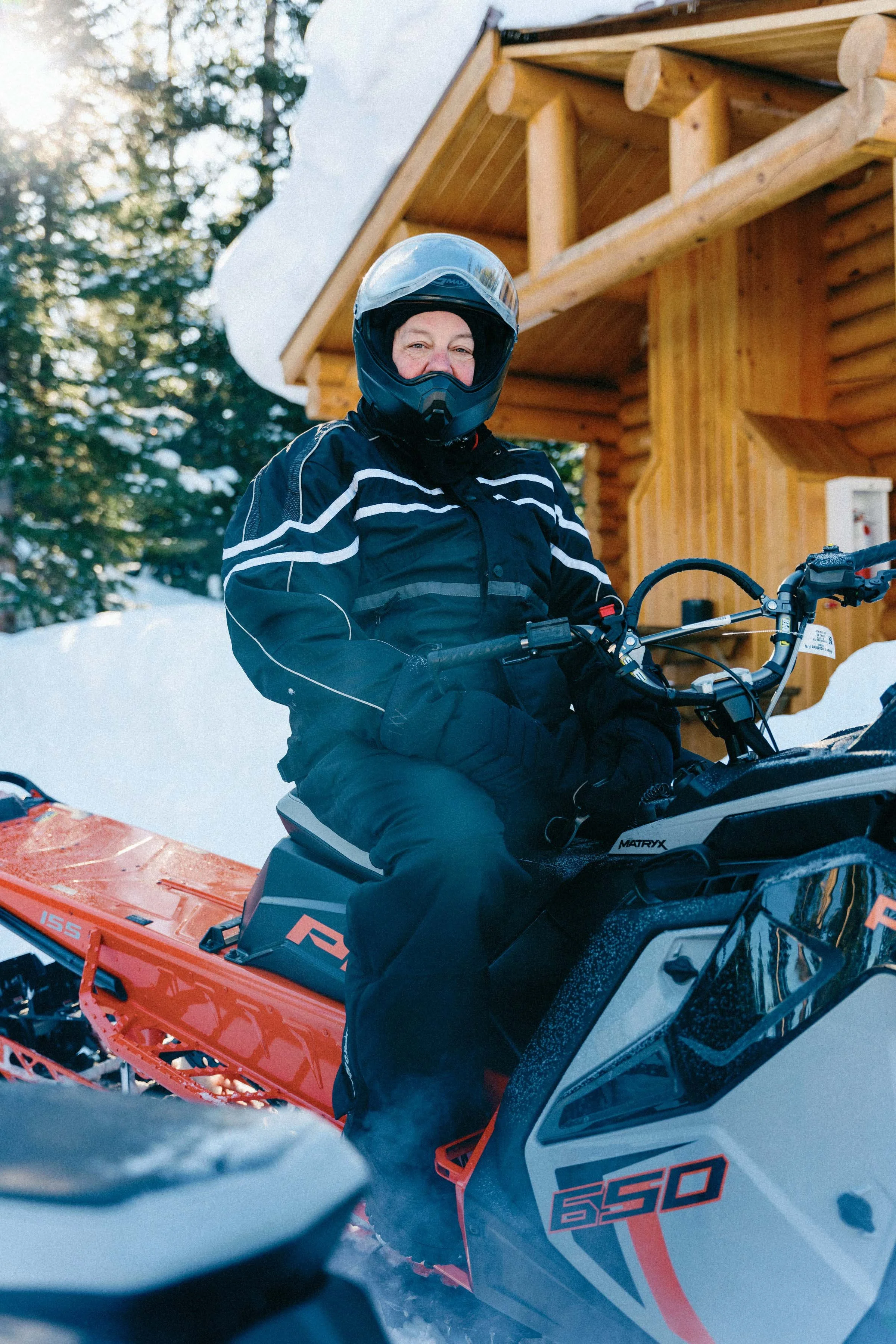 Person wearing a helmet and winter gear riding a snowmobile in a snowy outdoor setting with trees and a wooden cabin.