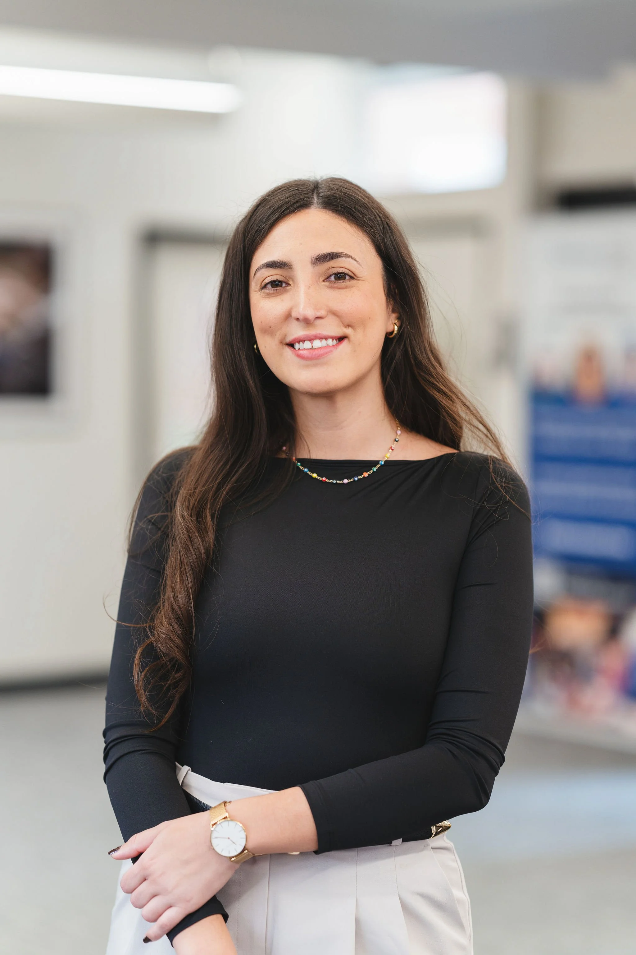 A young woman with long, dark hair and light skin, wearing a black long-sleeve top and white pants, standing indoors with a smile.