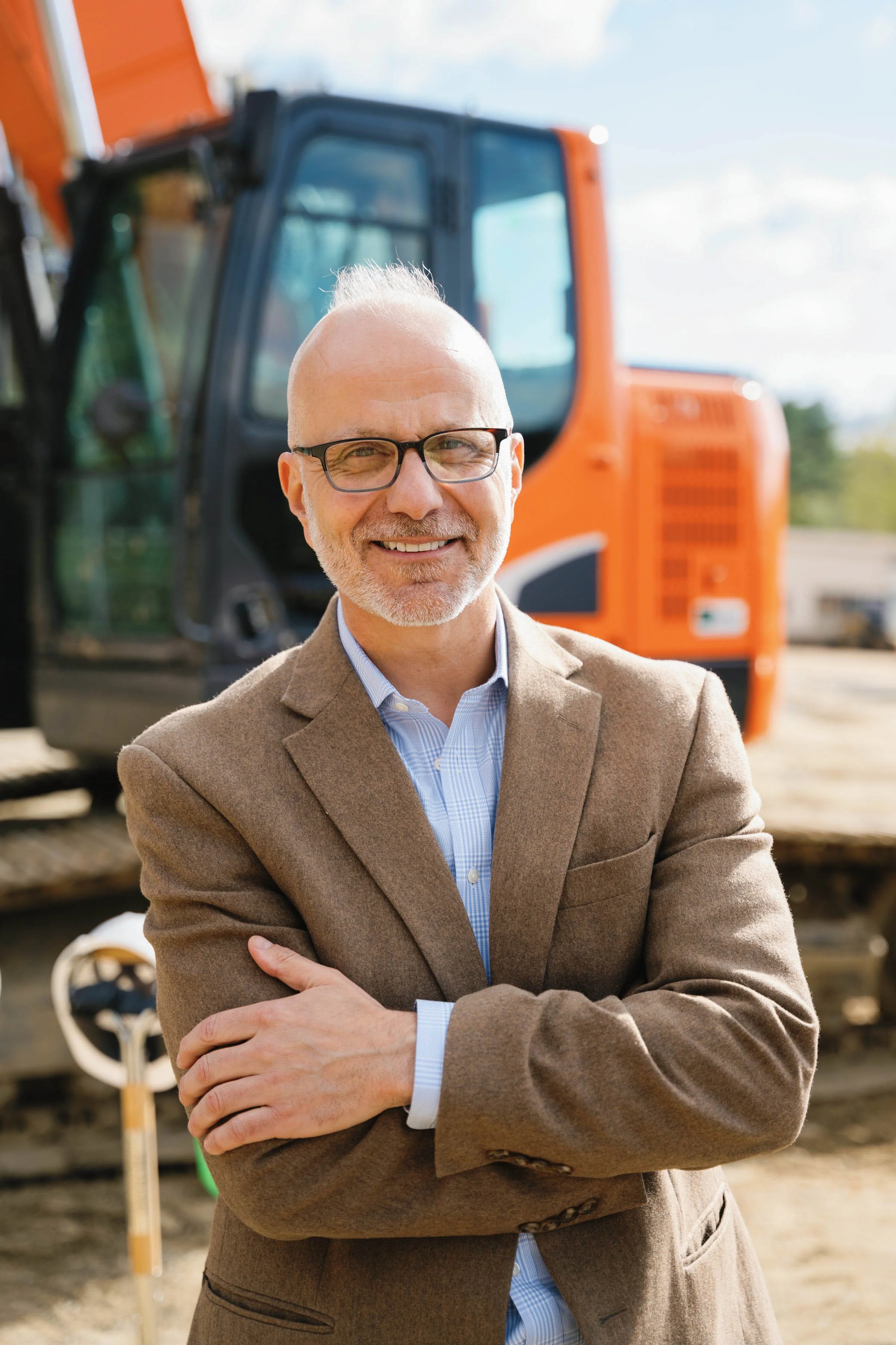 A smiling middle-aged man with glasses and a beard, dressed in a brown blazer and blue shirt, standing outdoors in front of construction equipment, including an orange excavator.