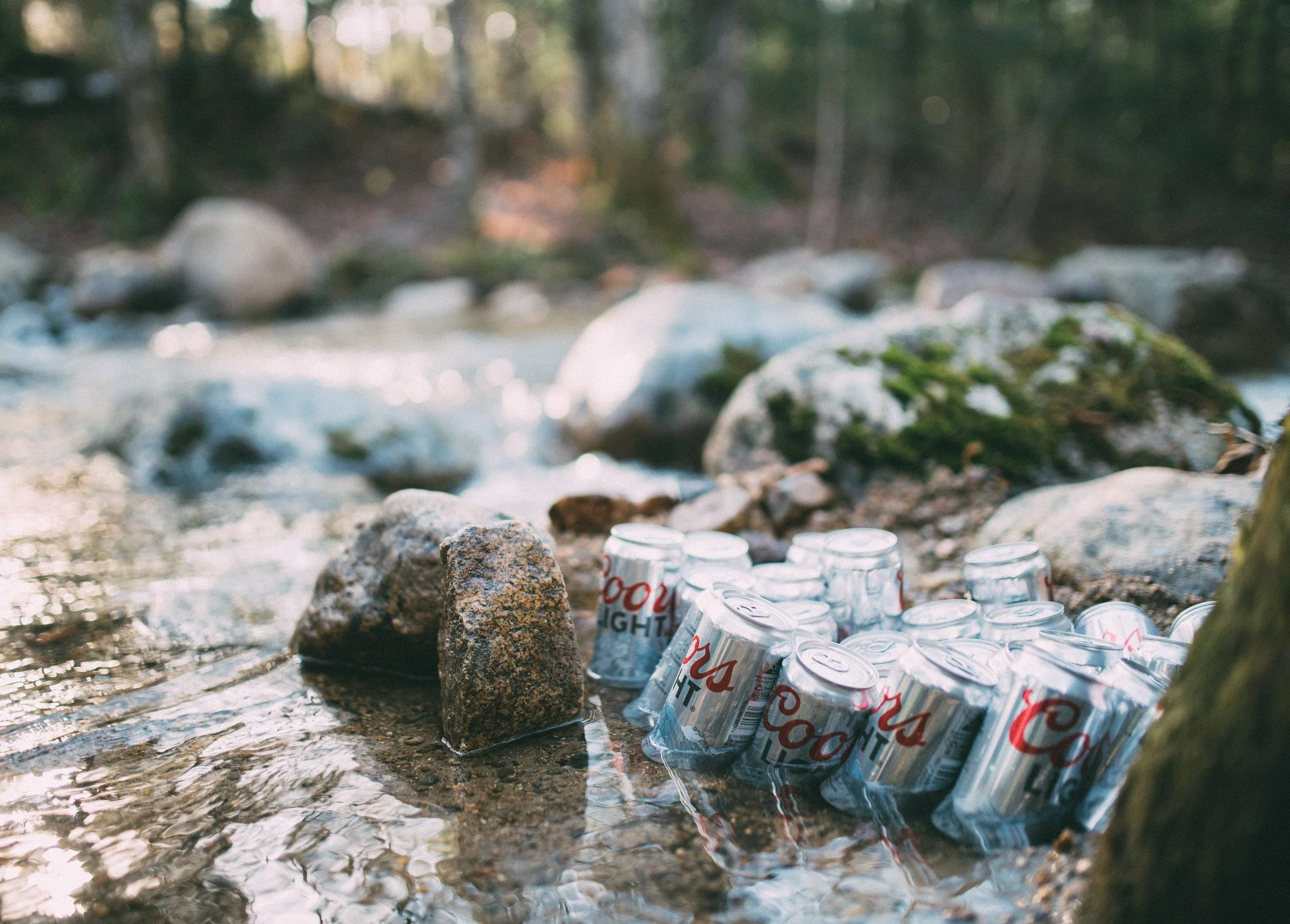 A collection of Coors Light beer cans partially submerged in a shallow creek with rocks and green moss, set in a forested outdoor environment.