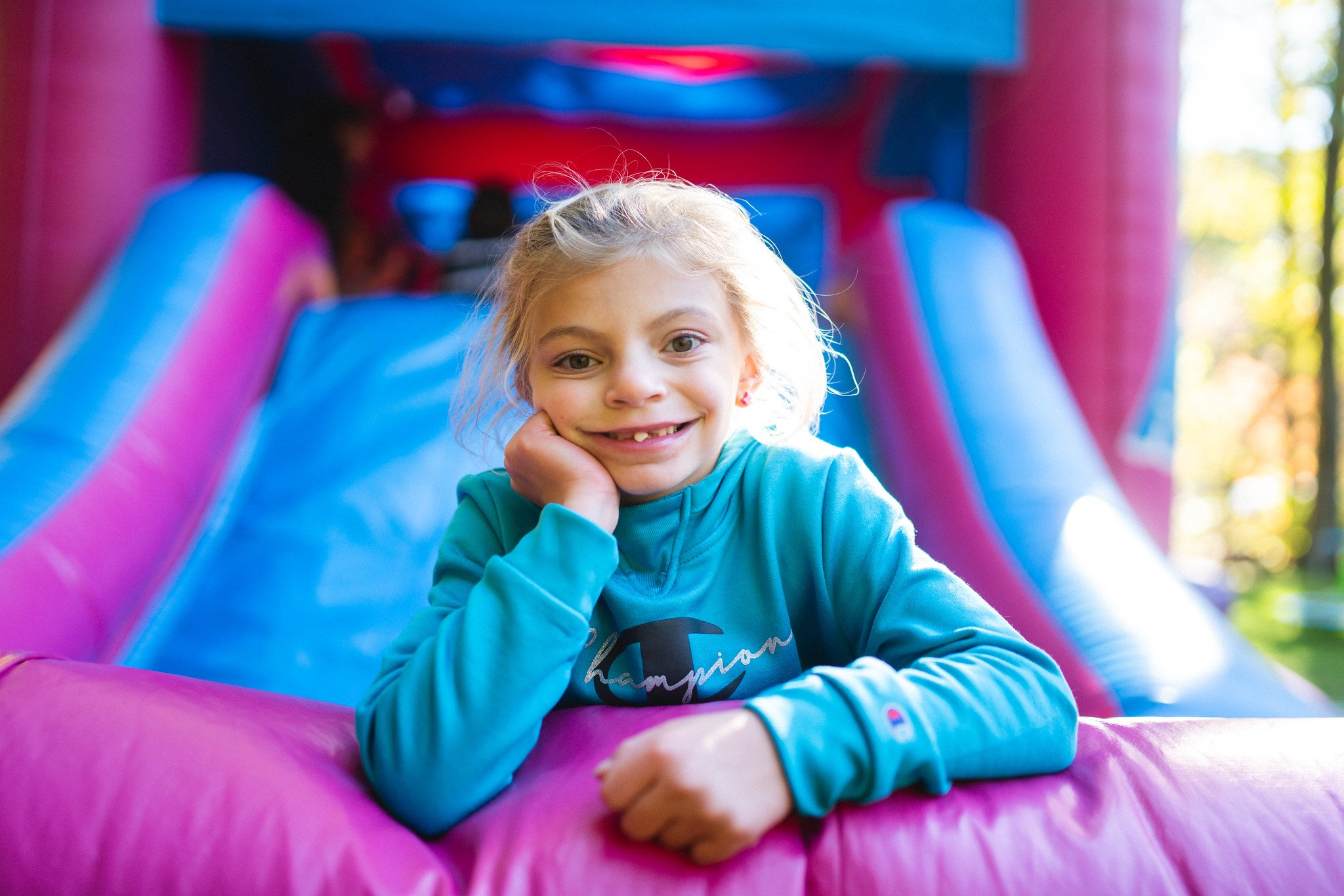 A young girl with blonde hair and a gap-tooth smile, lying on a pink padded surface of an inflatable bounce house or slide, resting her head on her hand, with a colorful play structure behind her.