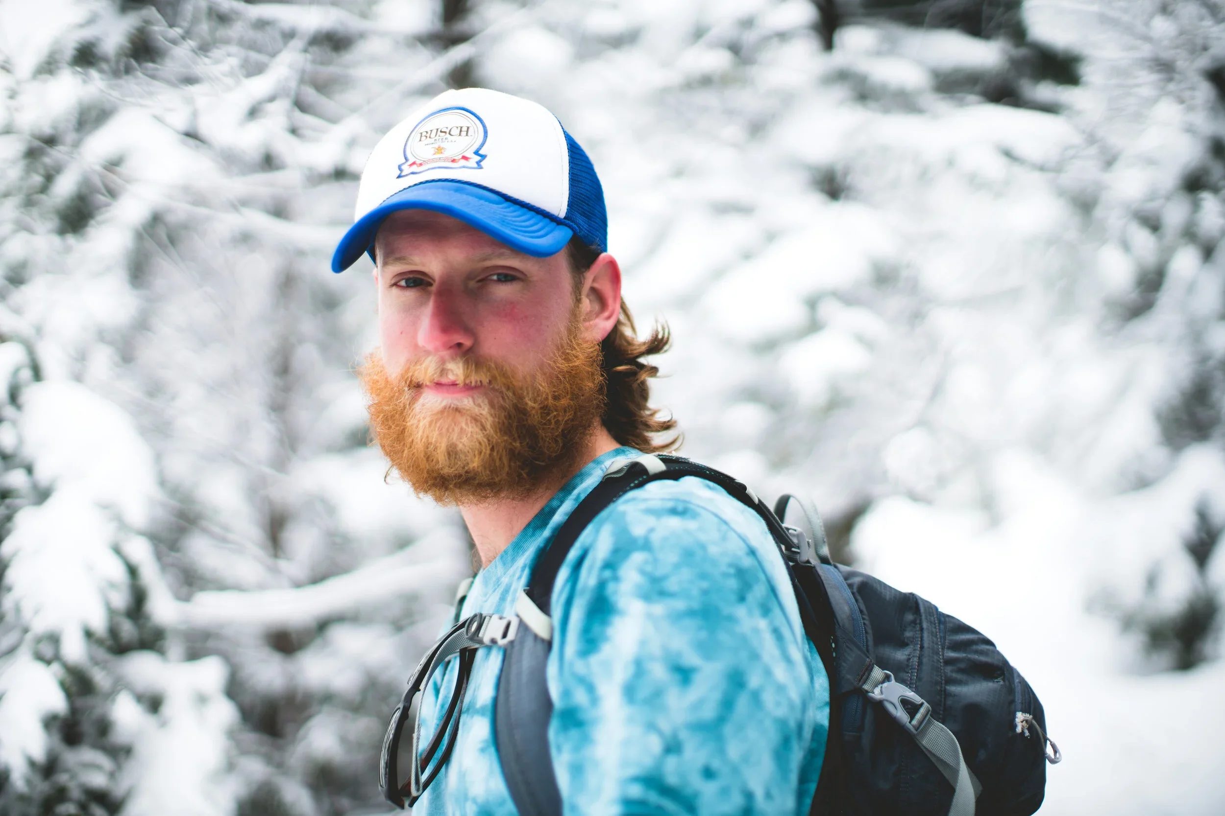 A man with a red beard and long hair wearing a blue and white cap, a blue tie-dye shirt, and a black backpack standing in a snowy forest.