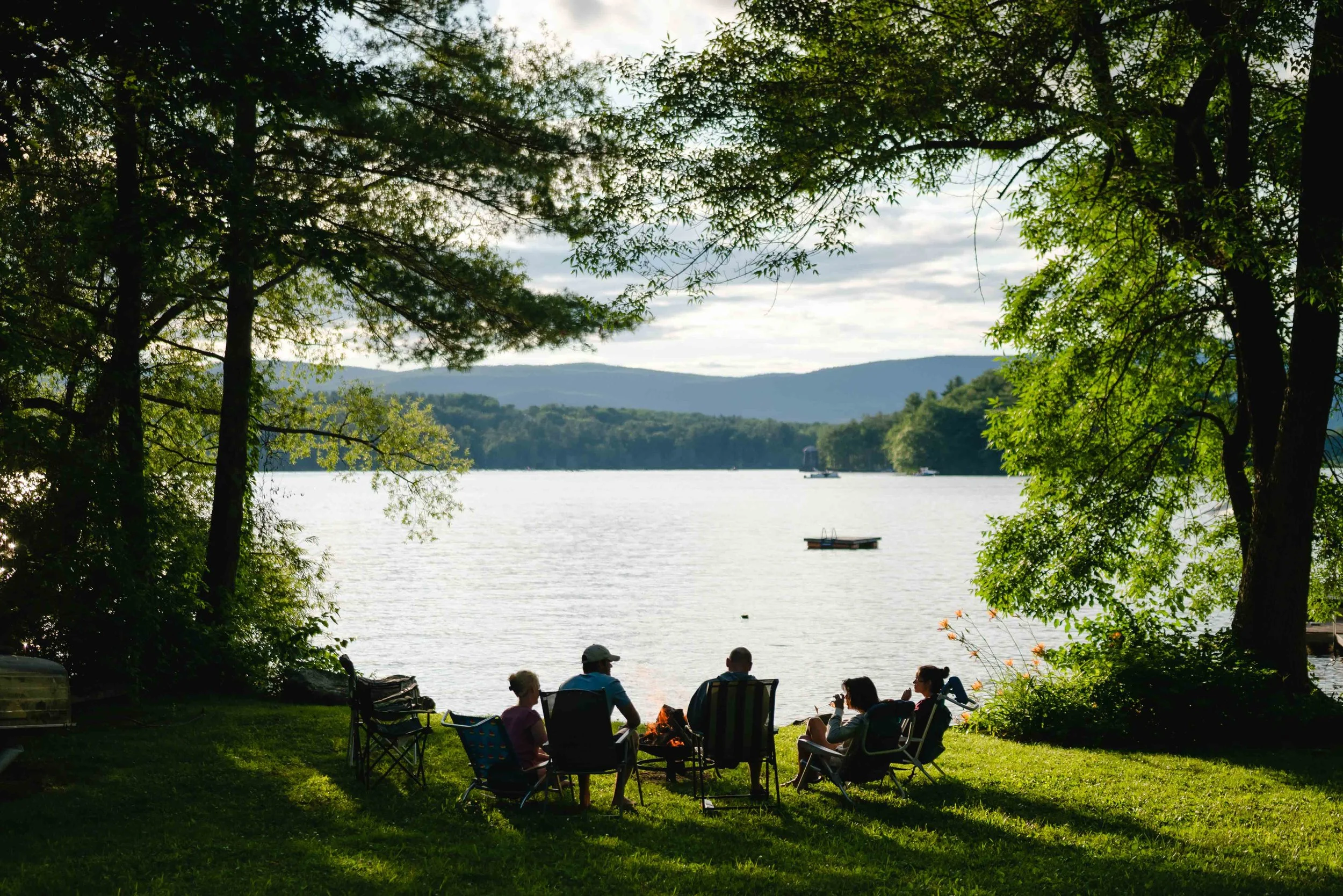 People sitting outdoors under trees near a lake, with mountains in the background.