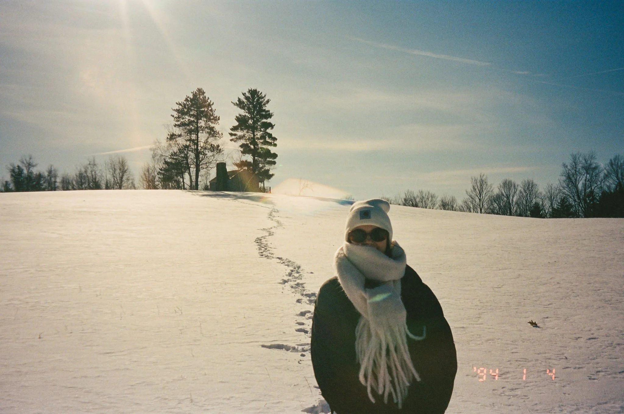 Person in winter clothing, including scarf, sunglasses, and a beanie, standing on snow-covered ground with a house and trees in the background on a sunny winter day.