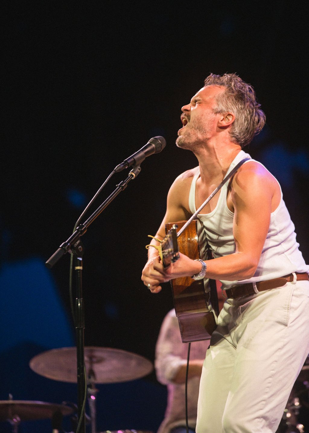 A man passionately singing and playing guitar on stage, wearing a white tank top and light-colored pants, against a dark background.