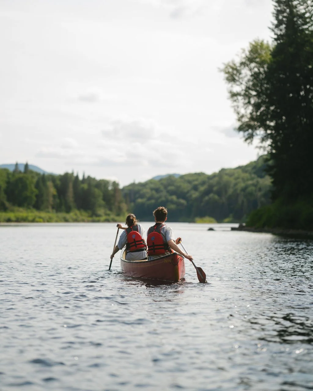 Two people in a red canoe paddling on a calm river surrounded by lush green trees and hills under a partly cloudy sky.