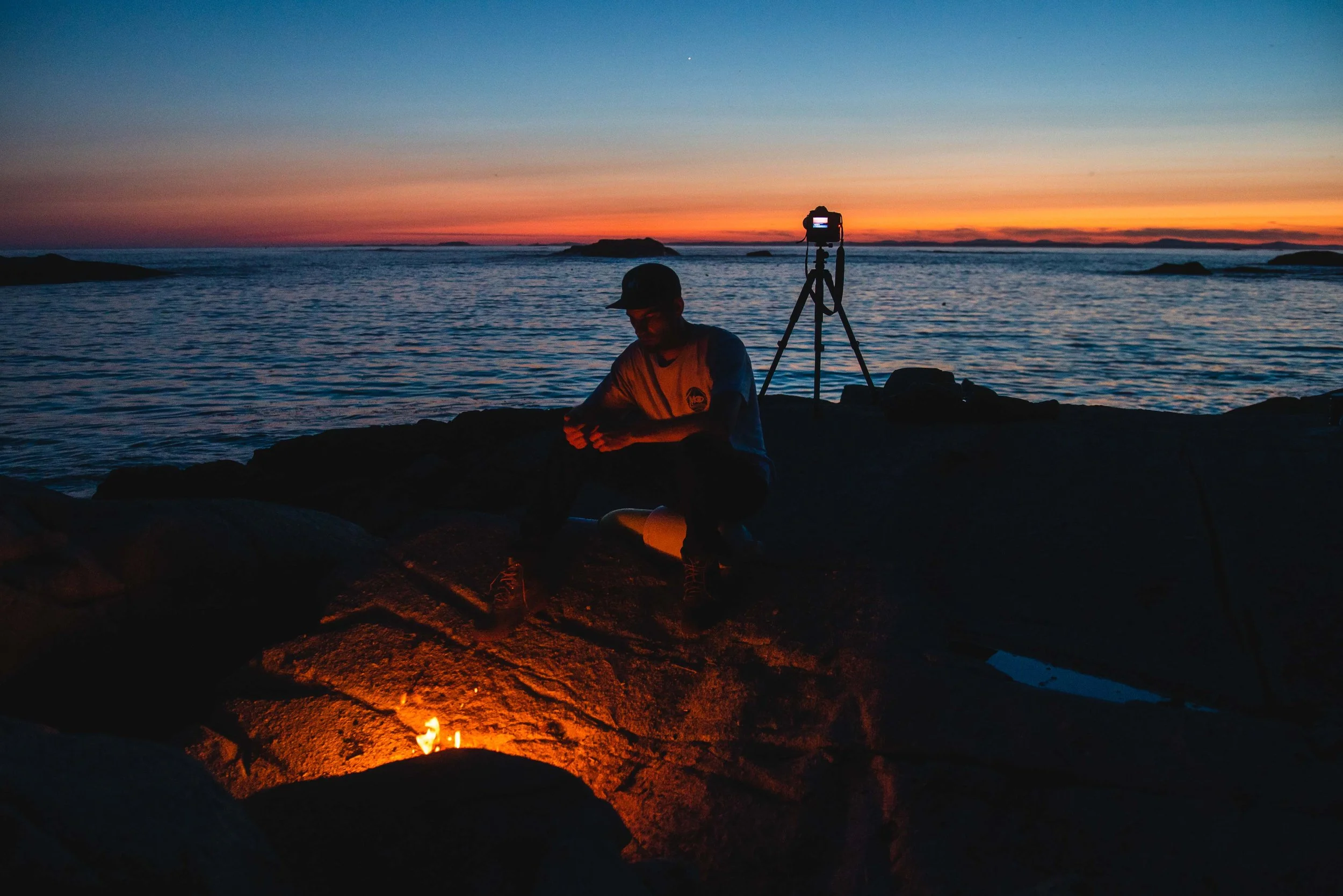 Person sitting on rocks by the ocean at sunset, with a camera on a tripod nearby, and a small fire burning in the foreground.