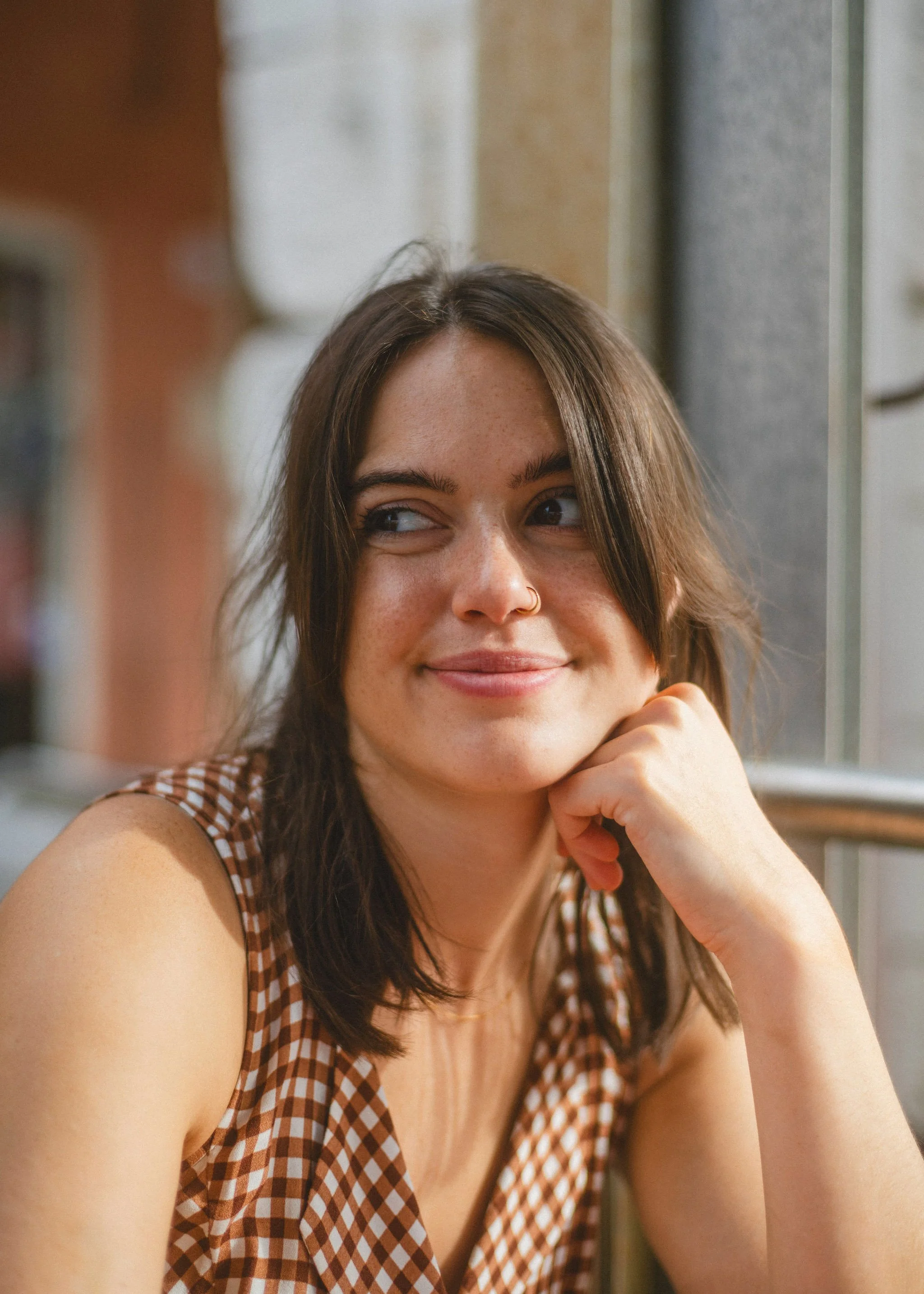 A woman with brown hair and a nose piercing sitting by a window, smiling and looking to the side.