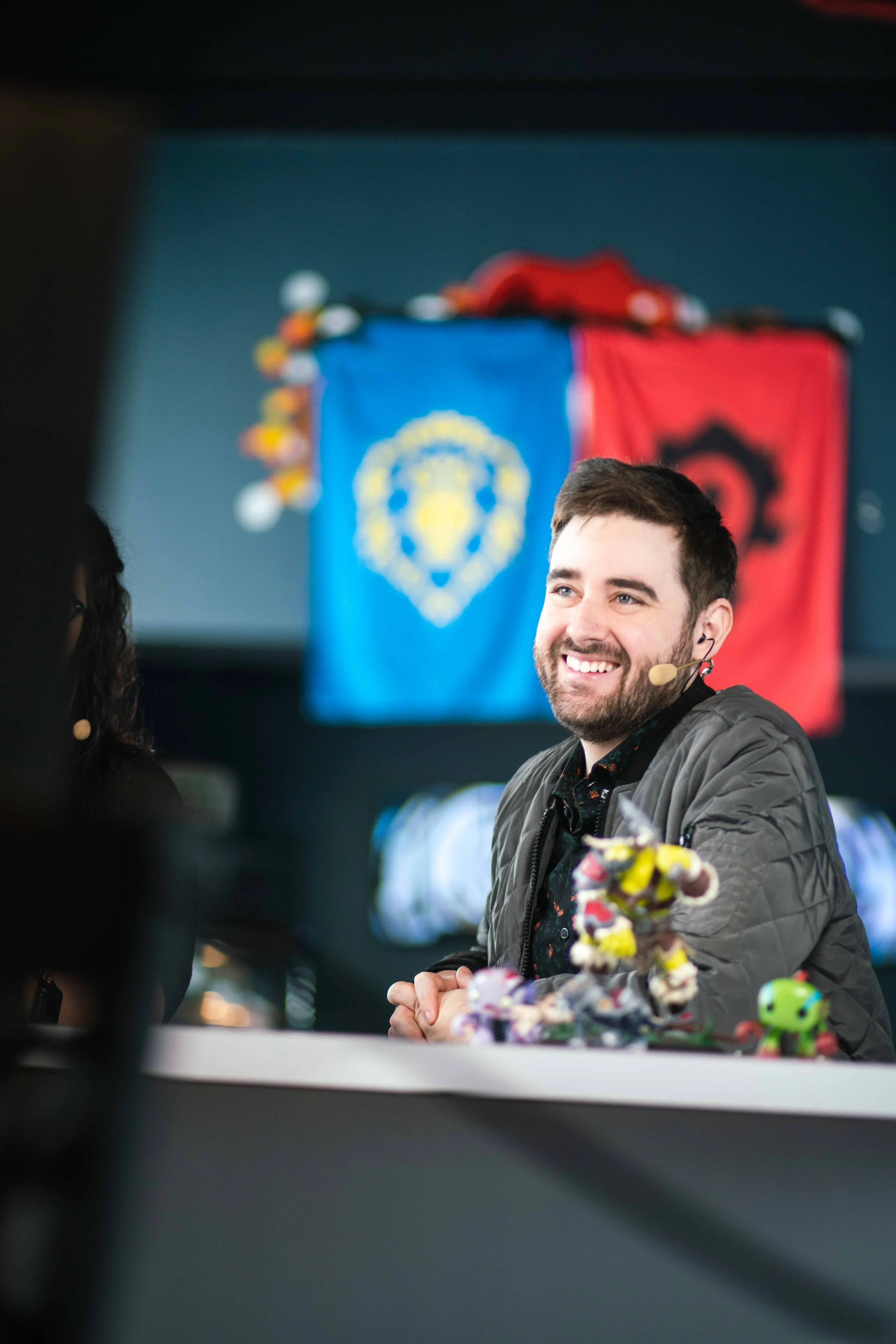 A man smiling with a headset microphone, sitting at a table with gaming figures, with a blue and red flag in the background.