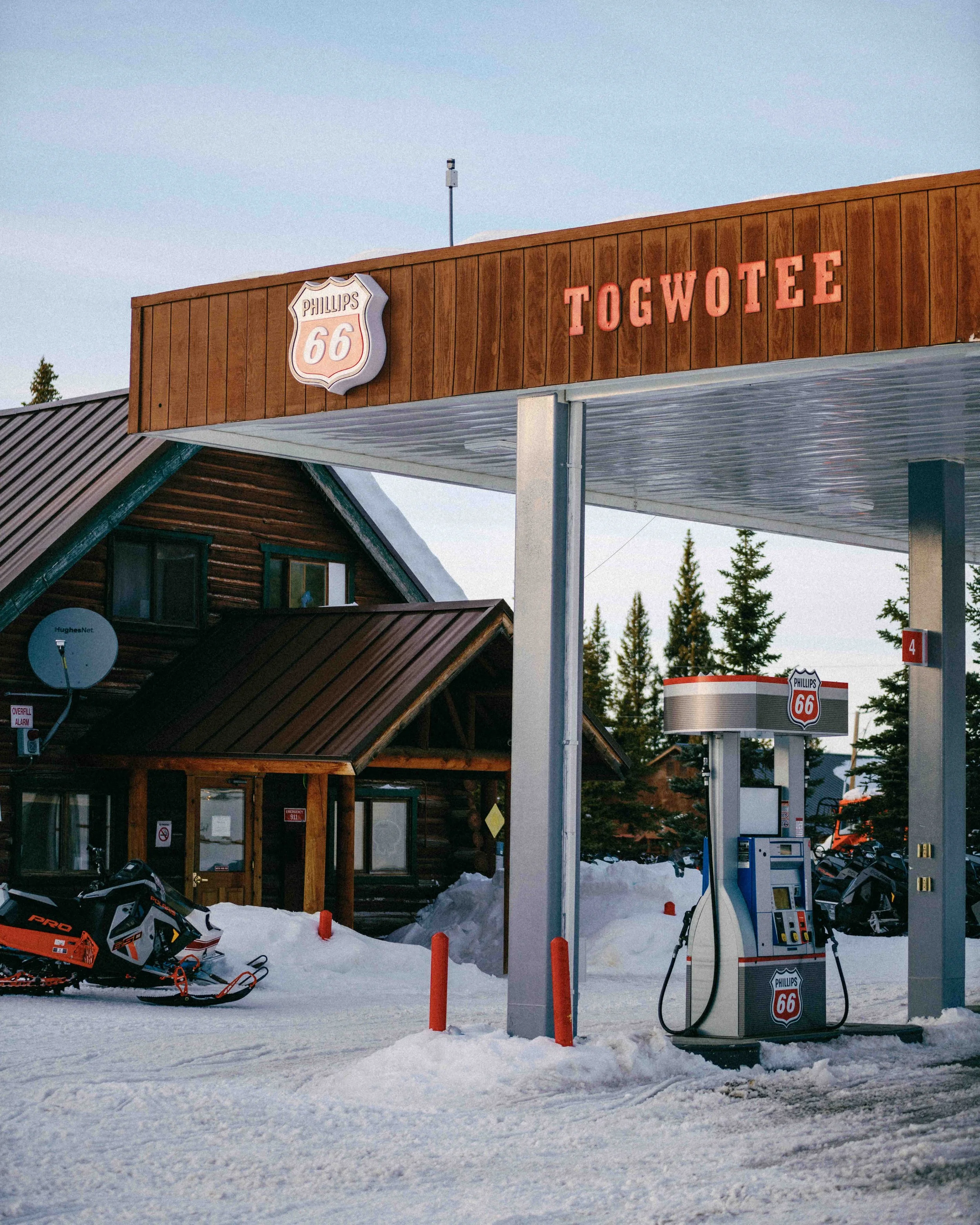 A gas station with a Phillips 66 sign, located in a snowy area with a snowmobile nearby. The station has a wooden canopy with the word 'TOGWOTEE' in red letters and a rustic wooden building in the background, with trees and a clear sky.