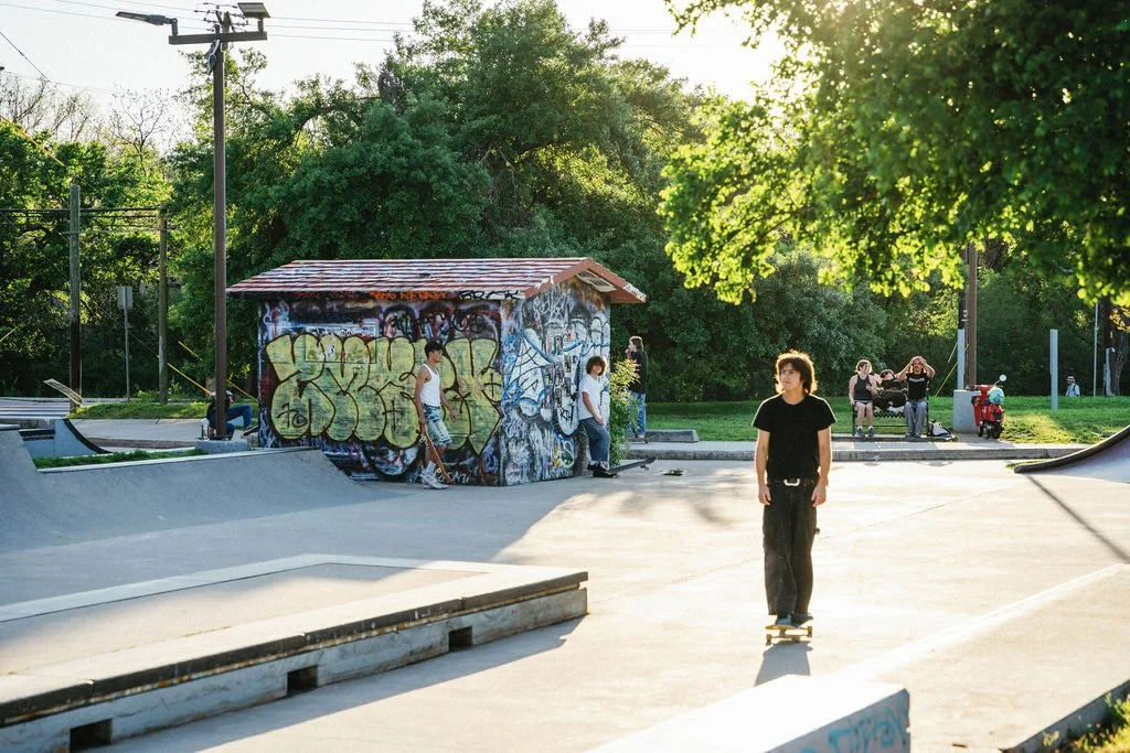 A skatepark with a young man skateboarding in the foreground, people in the background near graffiti-covered structures, and trees with sunlight filtering through.