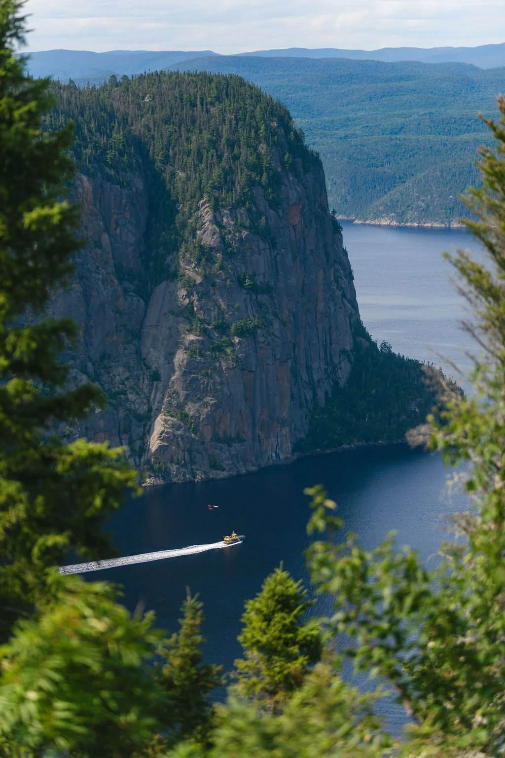 A boat sailing on a large blue lake with tall, forested cliffs on one side and mountains in the background, framed by green trees.