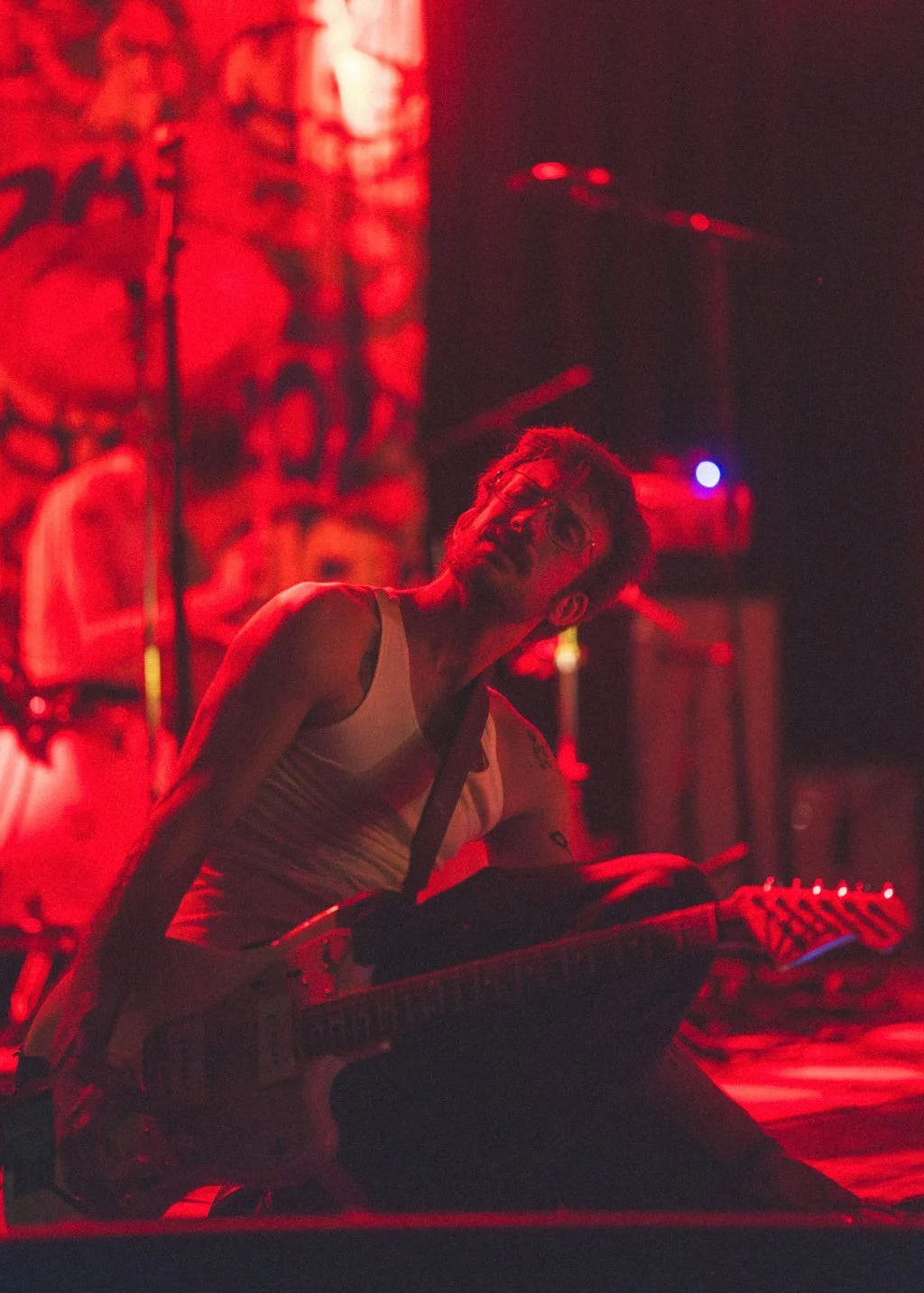The guitarist from model/actriz playing an electric guitar on stage, with red stage lighting and a backdrop featuring graffiti art.