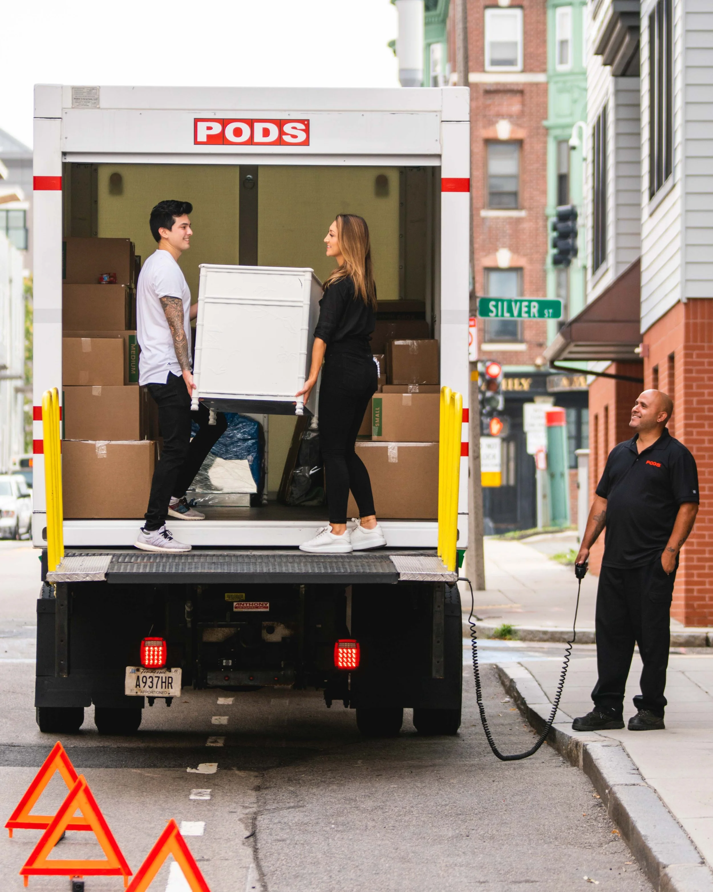 Two people loading moving boxes into a PODS unit on a city street, with the truck driver standing by.