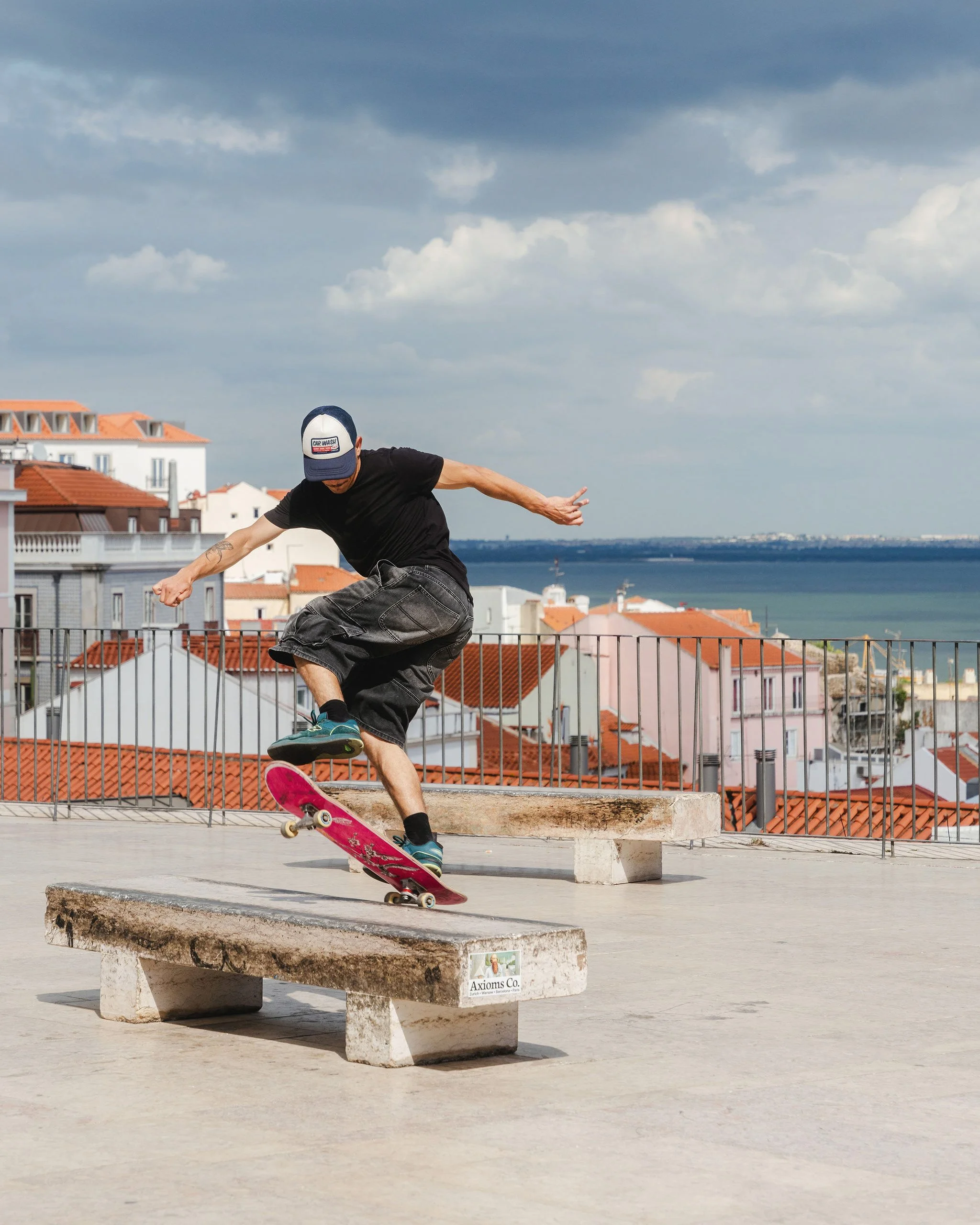 A skateboarder performing a trick on a concrete bench at a rooftop skate park with a cityscape and water in the background.