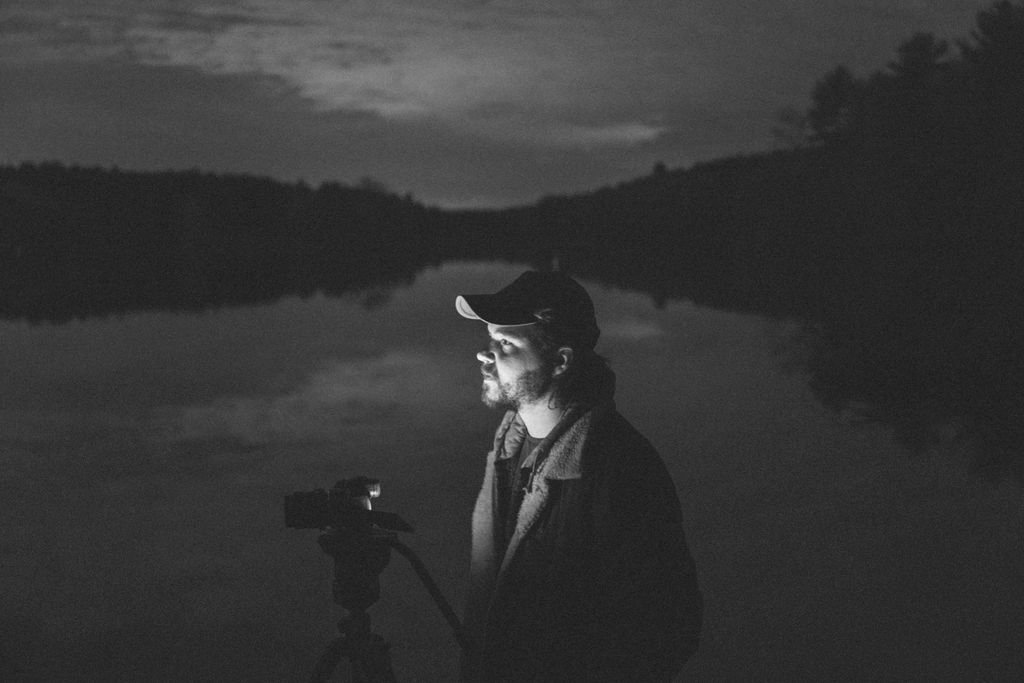 A man with a beard and long hair wearing a baseball cap and jacket, standing outdoors at night, looking at a camera on a tripod, with a body of water and hills in the background.