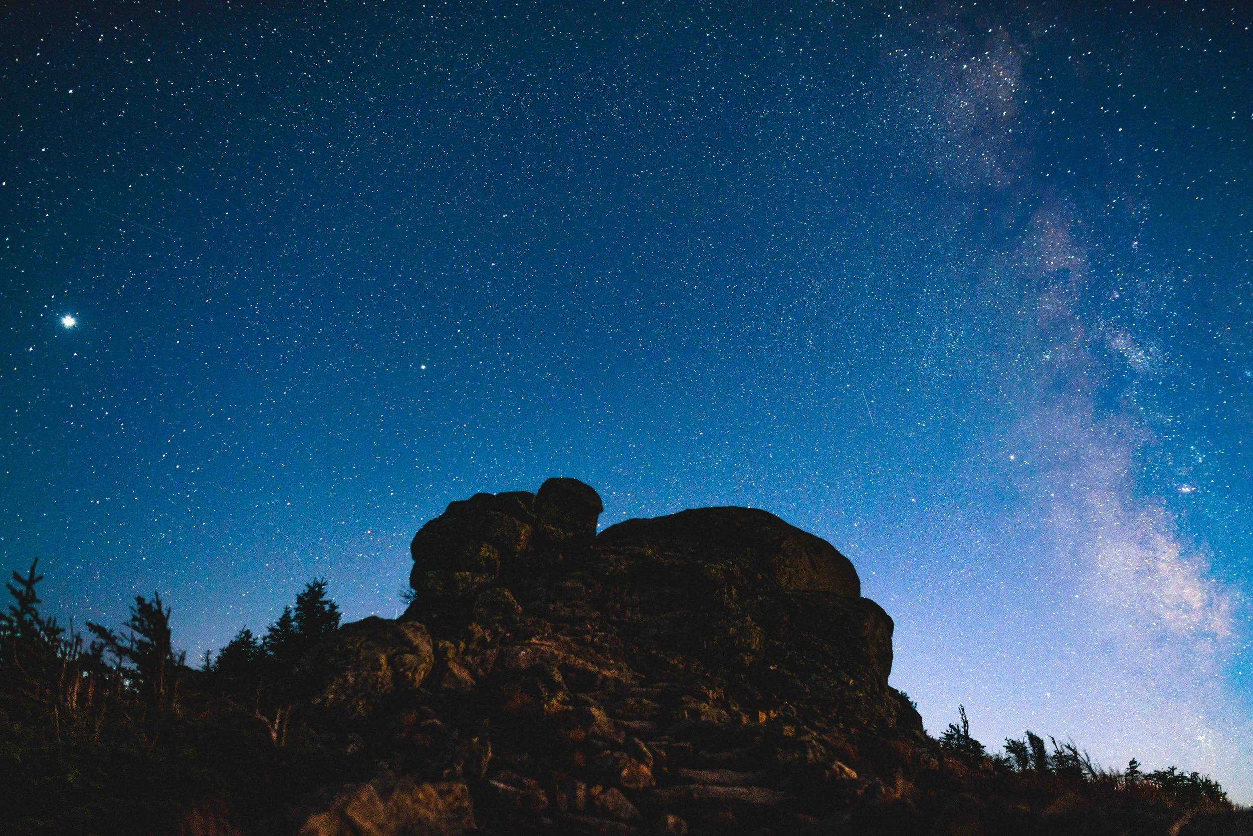 Night sky filled with stars and the Milky Way galaxy above a rocky hillside with trees in silhouette.