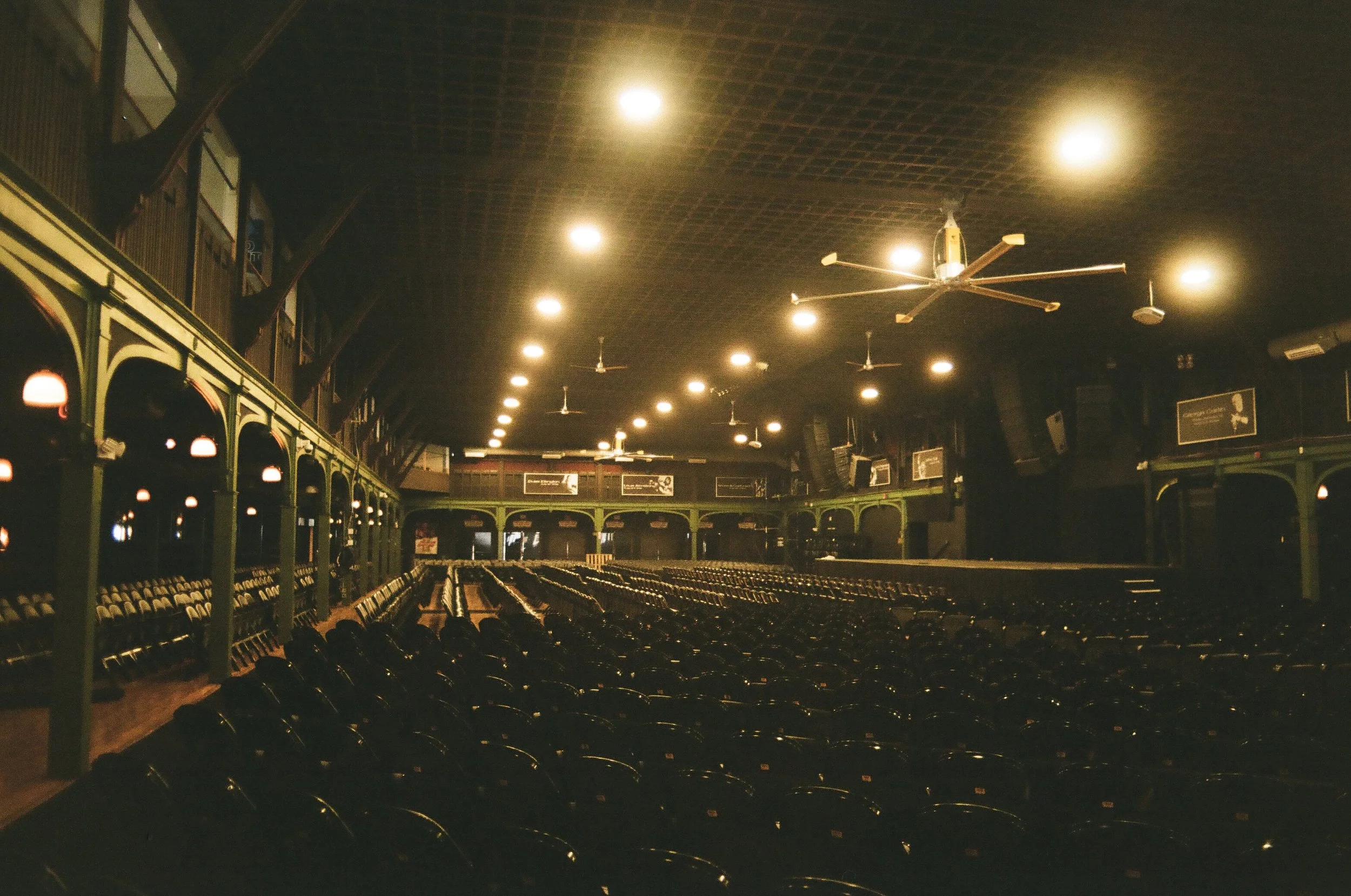 Empty indoor sports arena with rows of black seats, green balcony railings, multiple ceiling fans, and dim lighting.