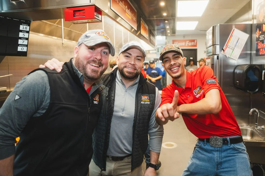 Three men smiling and posing inside a fast food restaurant, with one man pointing at the camera, wearing a red uniform with a 'Cane's' logo.