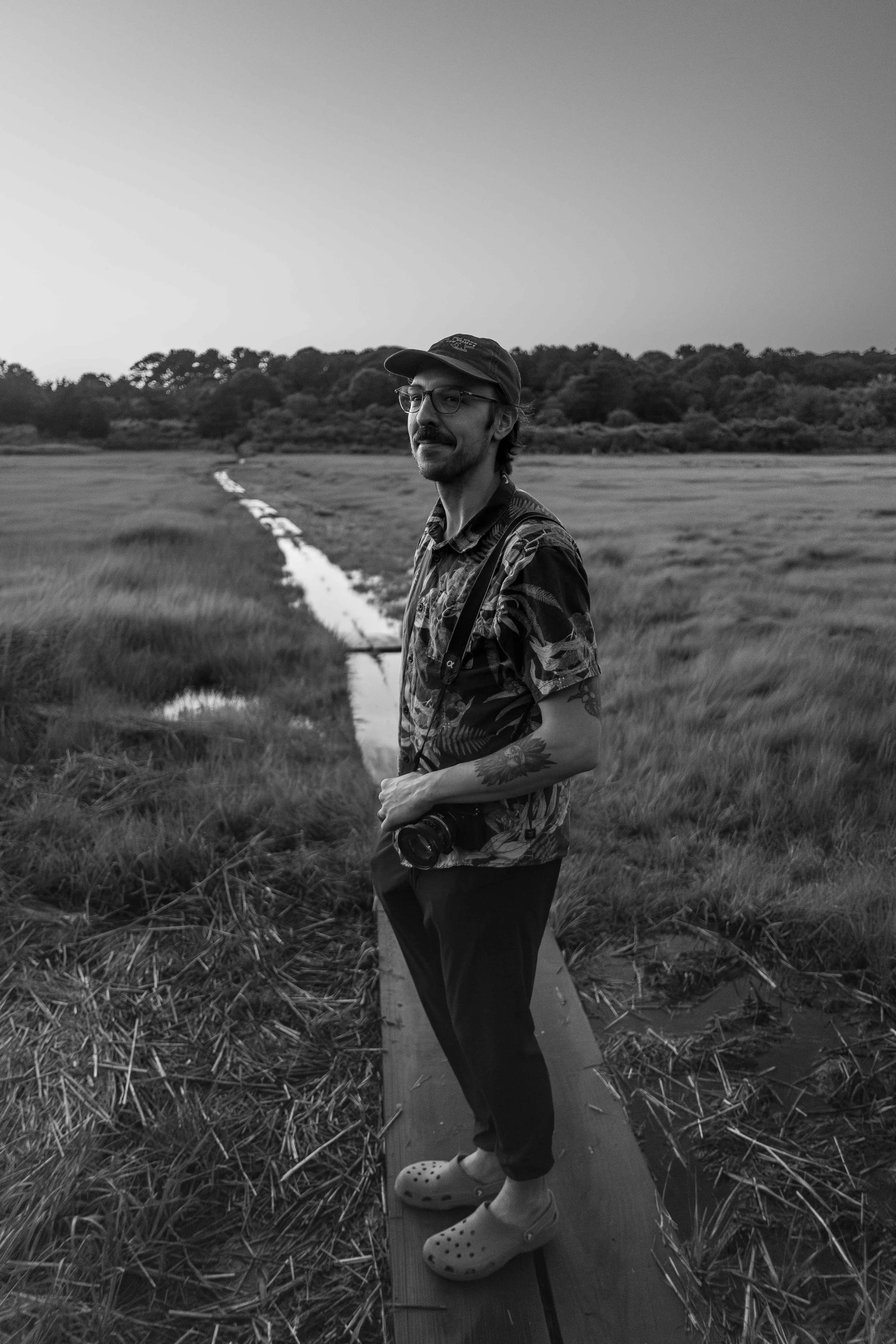A man standing on a wooden walkway in a wetland. He is wearing a hat, glasses, a patterned shirt, black pants, and Crocs. He has a camera hanging around his neck and a tattoo on his right arm. In the background, there is a small stream and a landscape of trees and grass.