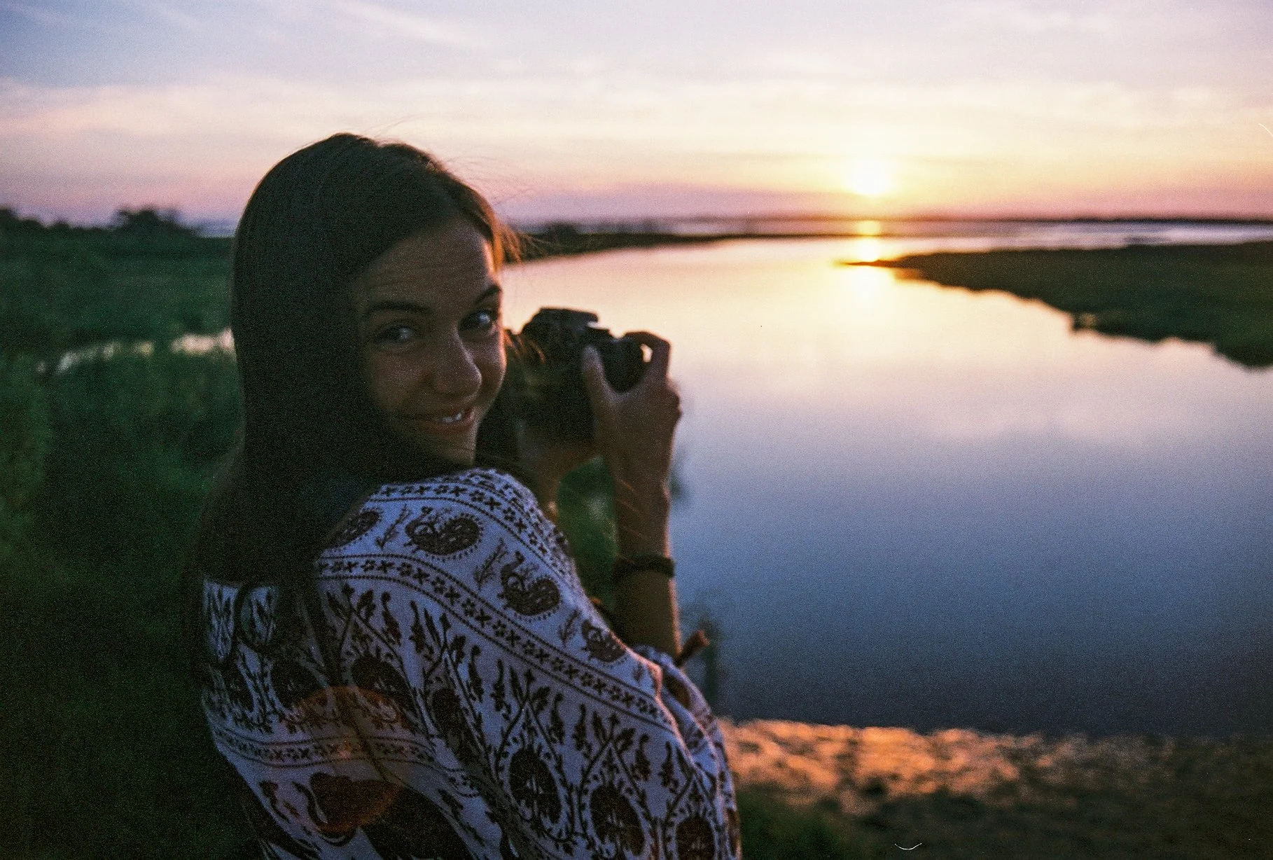 A woman with a patterned shawl, smiling, holding a camera, sitting by a river during sunset.