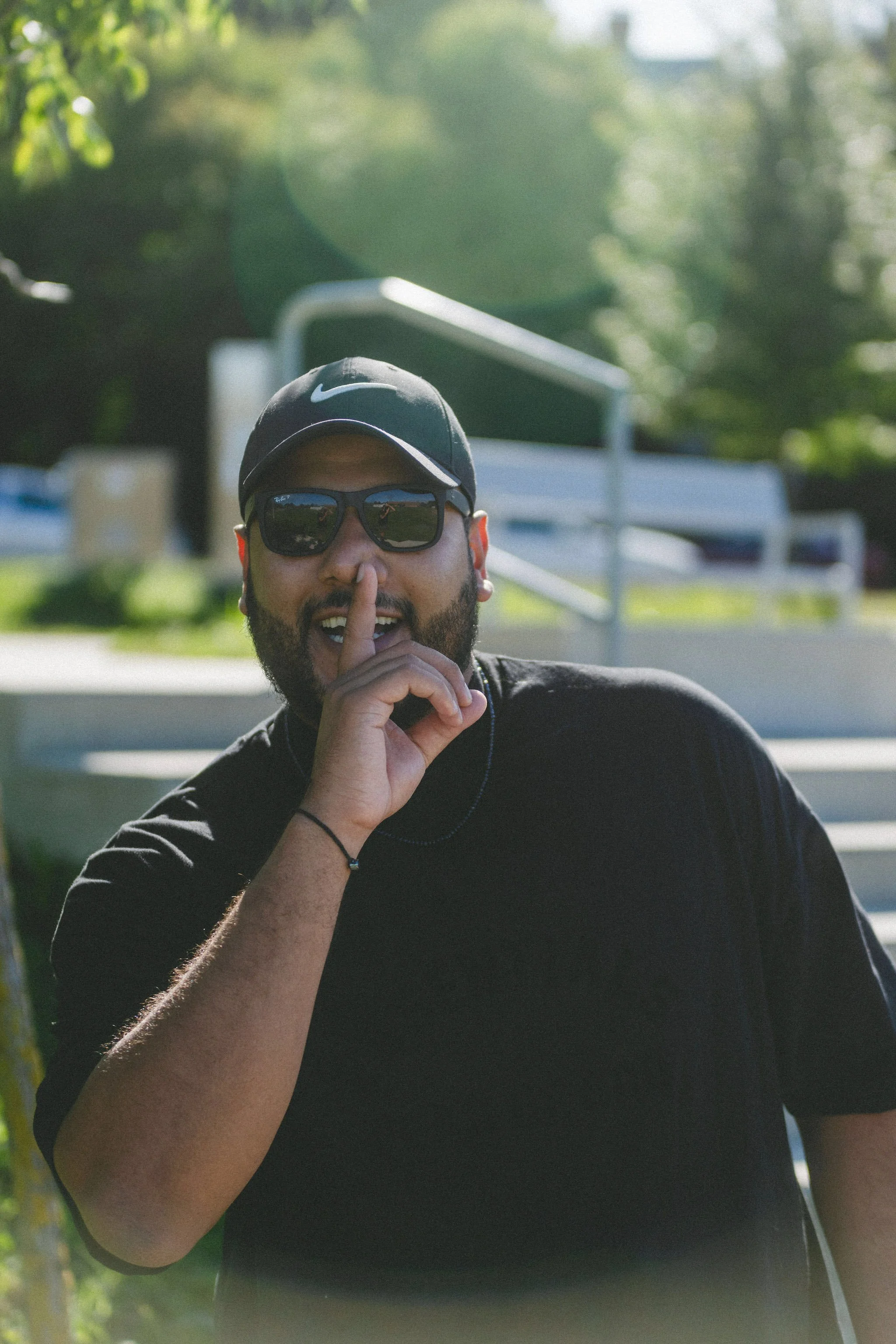 A man wearing sunglasses and a black cap with a Nike logo making a gesture by placing his finger on his lips, outdoors on a sunny day.
