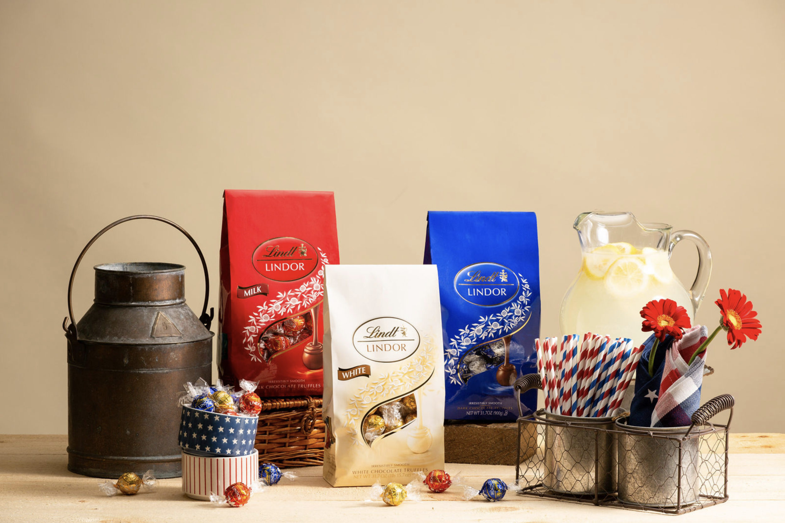 Assorted Lindt Lindor chocolate bags in red, blue, and white with a metal ice cream tub filled with red, white, and blue striped straws and napkins, a glass pitcher of lemonade with lemon slices, a small blue and white star-patterned tin of chocolate