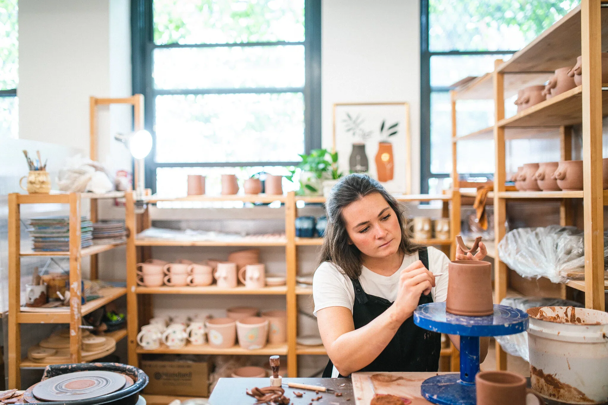A woman is pottery making in a ceramics studio surrounded by shelves of pottery and tools.