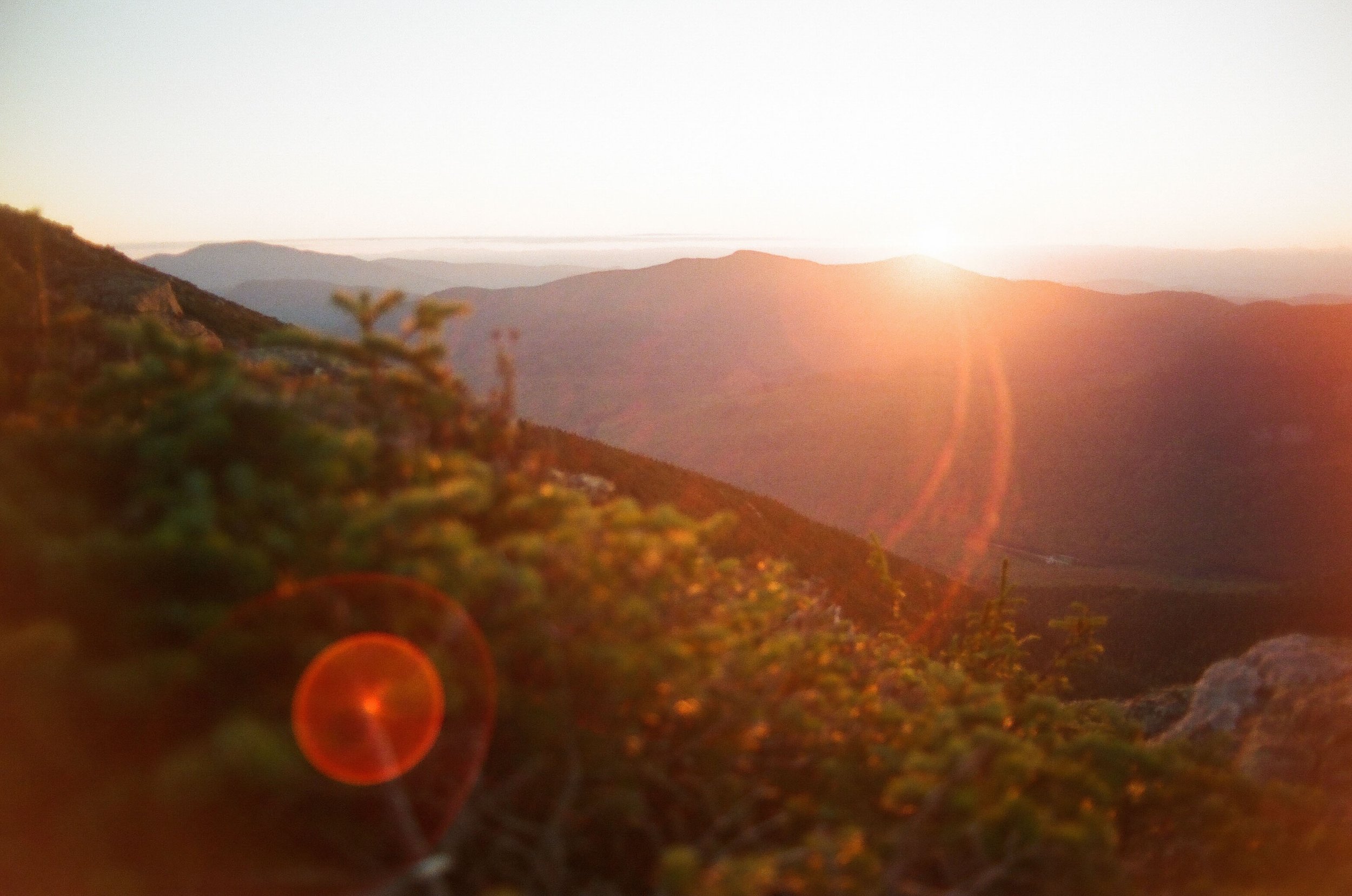 Sunset over a mountainous landscape with a warm glow and lens flare, including trees and rolling hills.