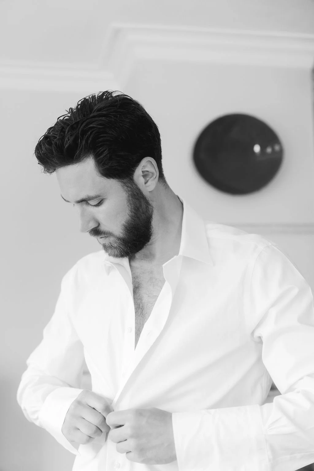 A man with dark hair and a beard, wearing a white shirt, buttoning his shirt in a room with plain walls and a circular black wall clock.