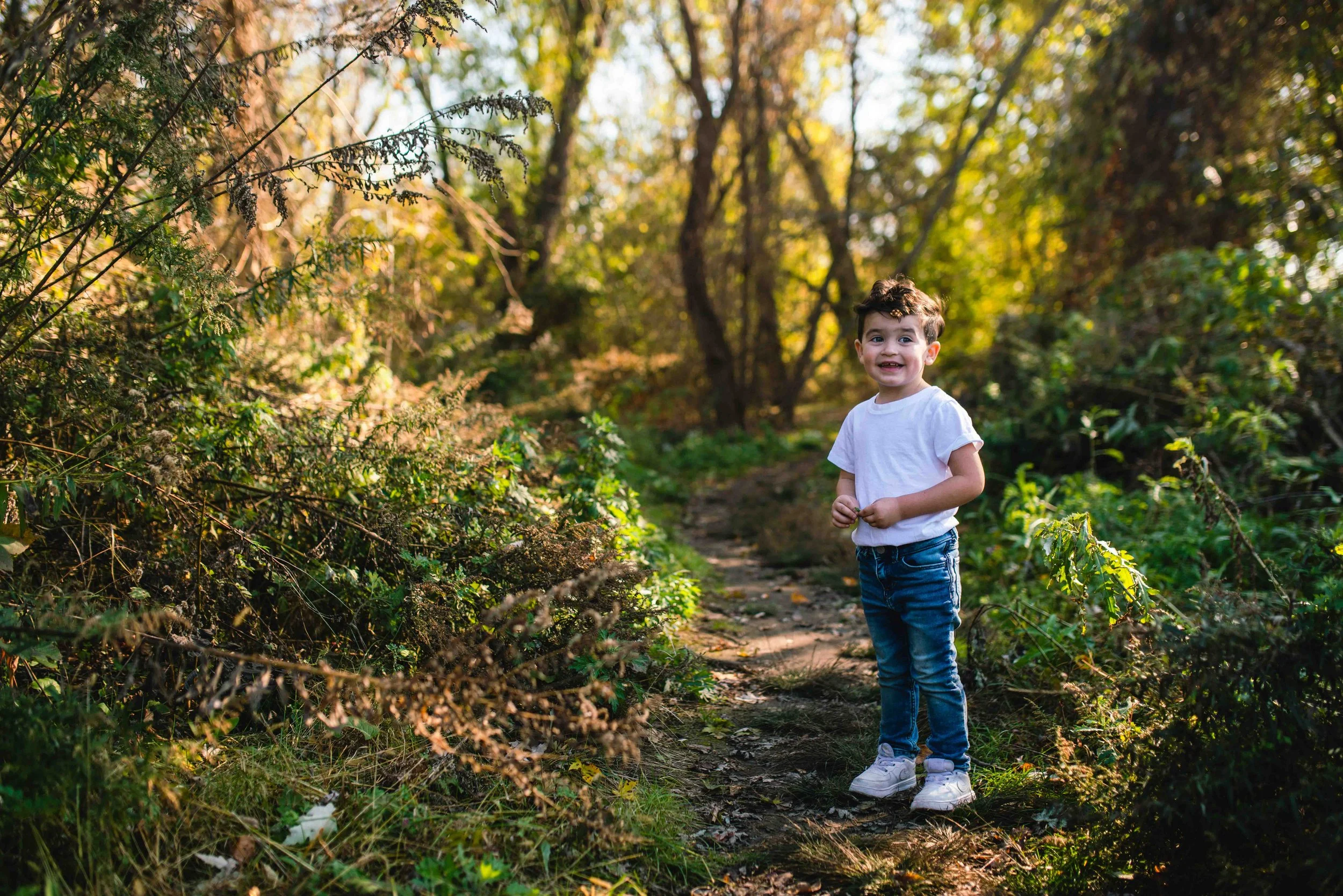 A young boy standing on a dirt path in a lush wooded area during autumn, smiling and looking at the camera.