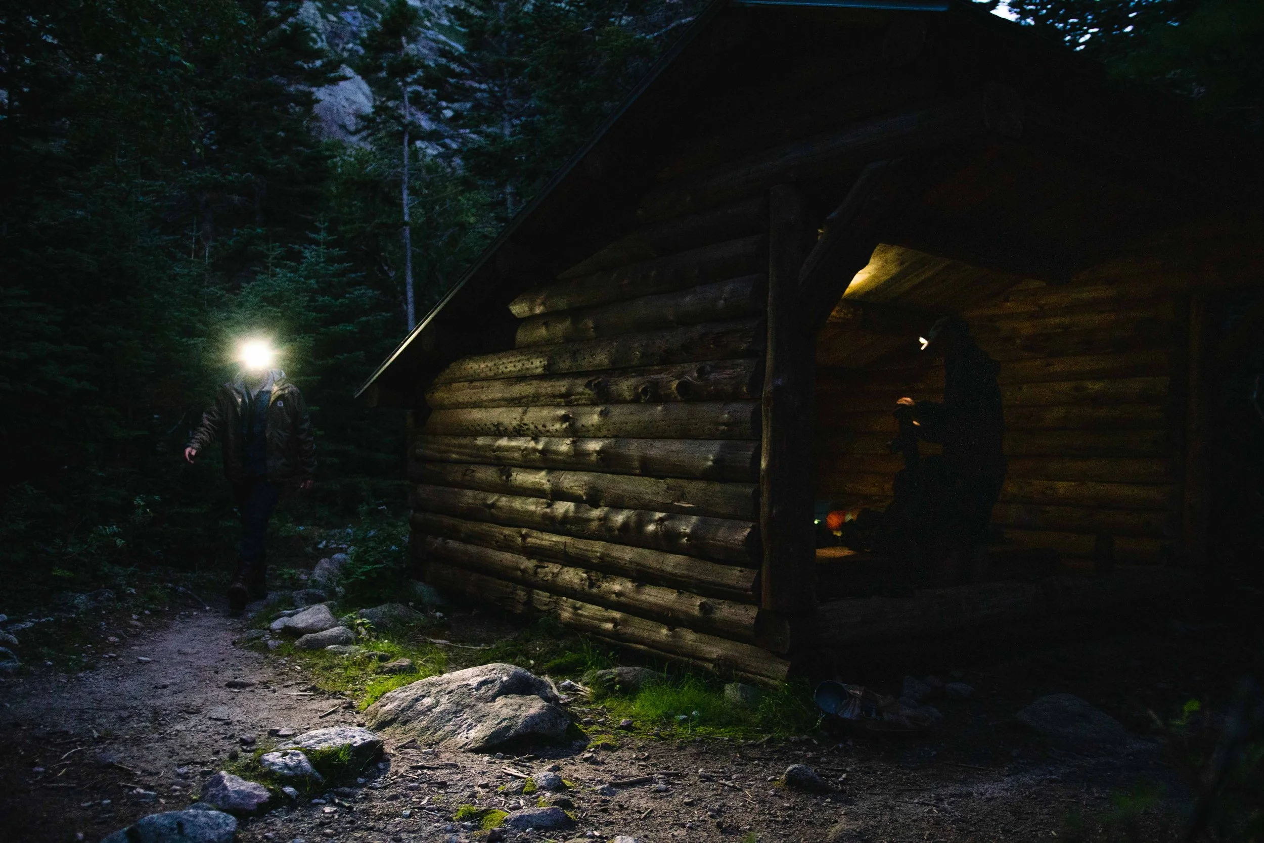 A person walking on a rocky trail at night with a headlamp on, approaching a wooden cabin in a forest.