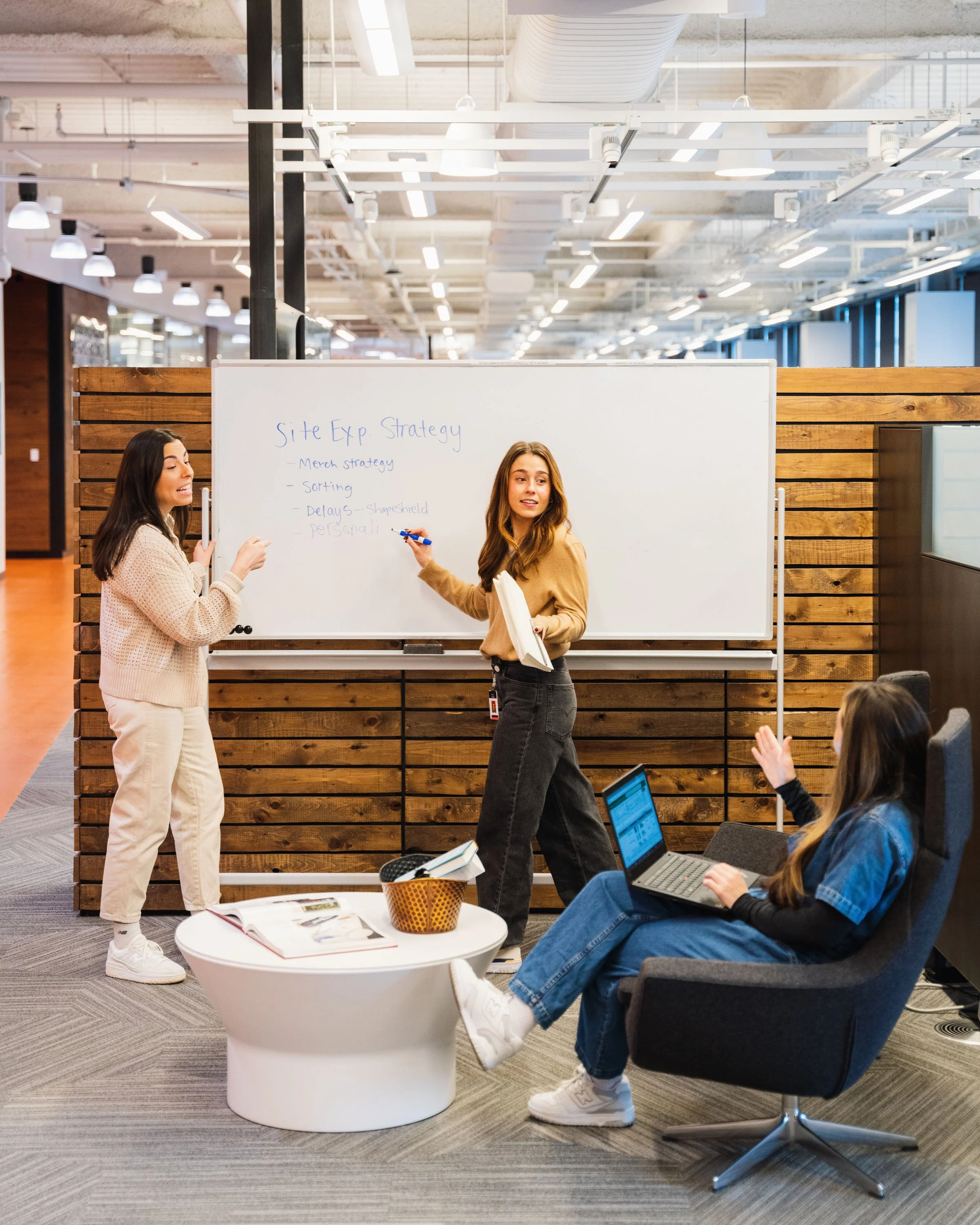 Three women in a modern office space having a discussion, with one woman standing and pointing at a whiteboard with strategy notes, and one woman sitting in a chair with a laptop, while another woman stands nearby.