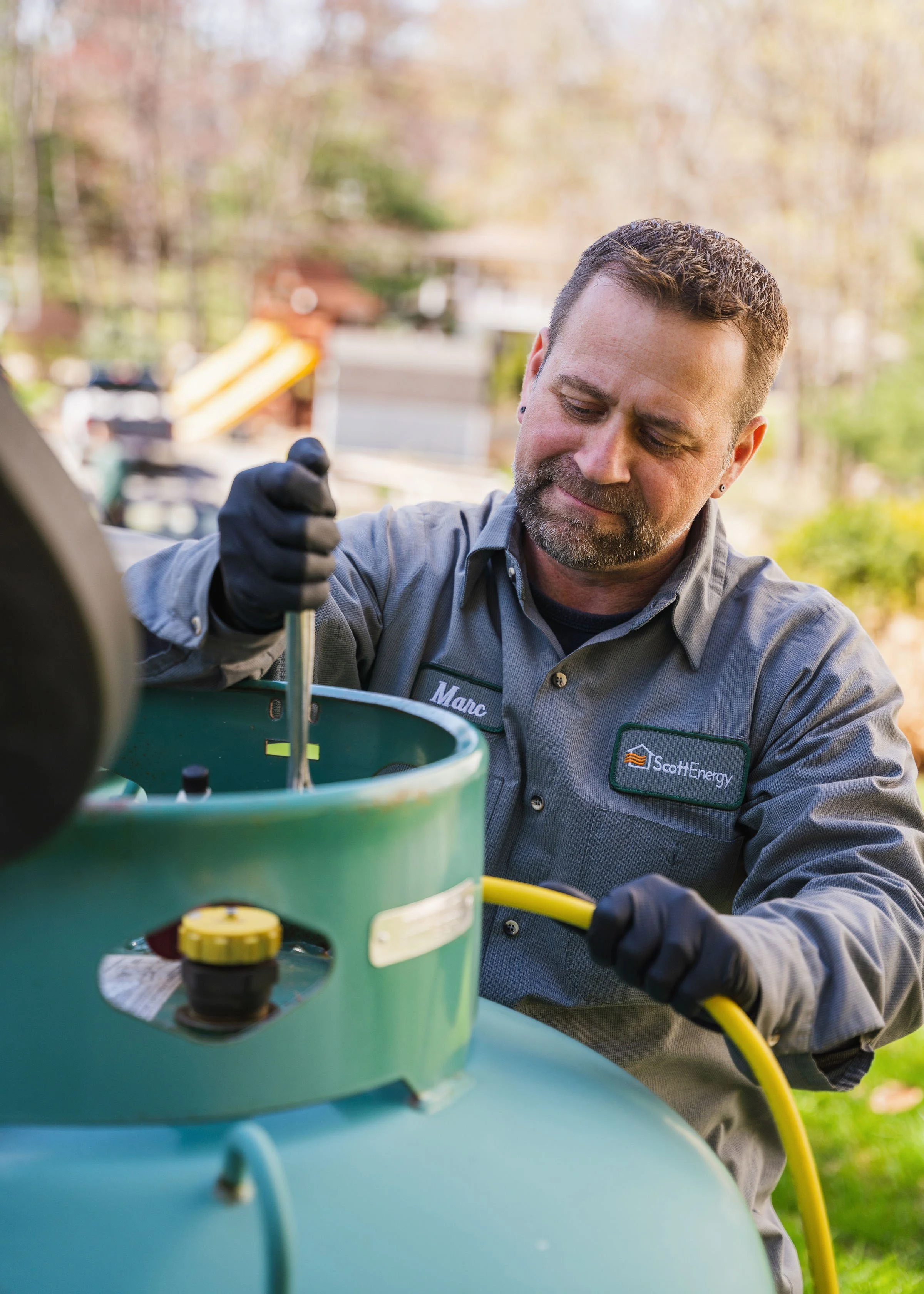 Man in work shirt and gloves working on a piece of equipment outdoors with trees in the background.