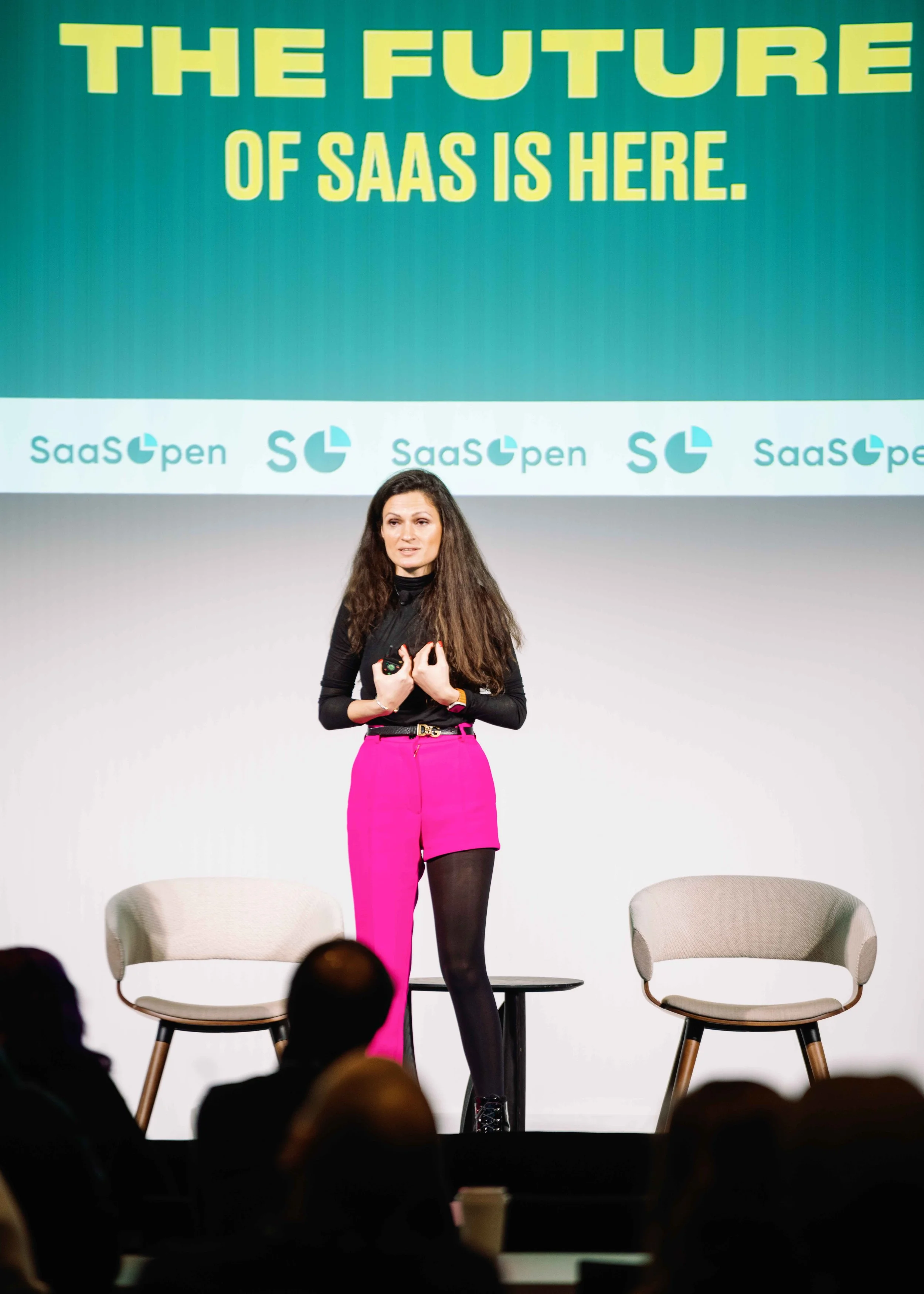 A woman with long brown hair, wearing a black top, pink pants, and black tights, is standing on stage speaking at SaaS open event. Two beige chairs are behind her, and an audience is visible at the bottom. The background features a large screen with 