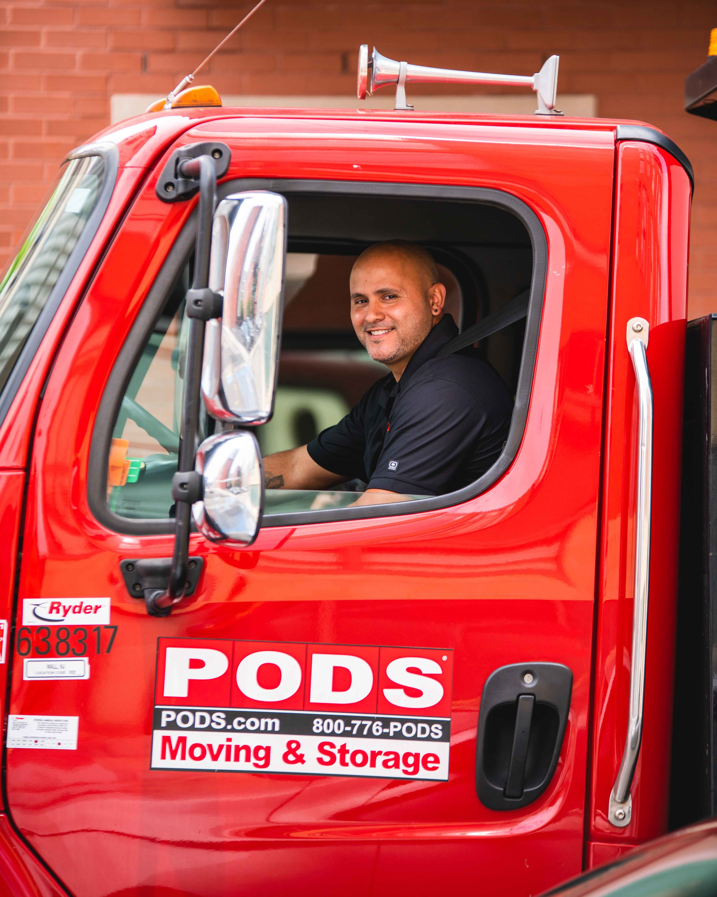 A person sitting in the driver's seat of a red moving truck. The truck has the company logo 'PODS' on the door along with contact information and the words 'Moving & Storage'. The person is smiling and looking out the open window.