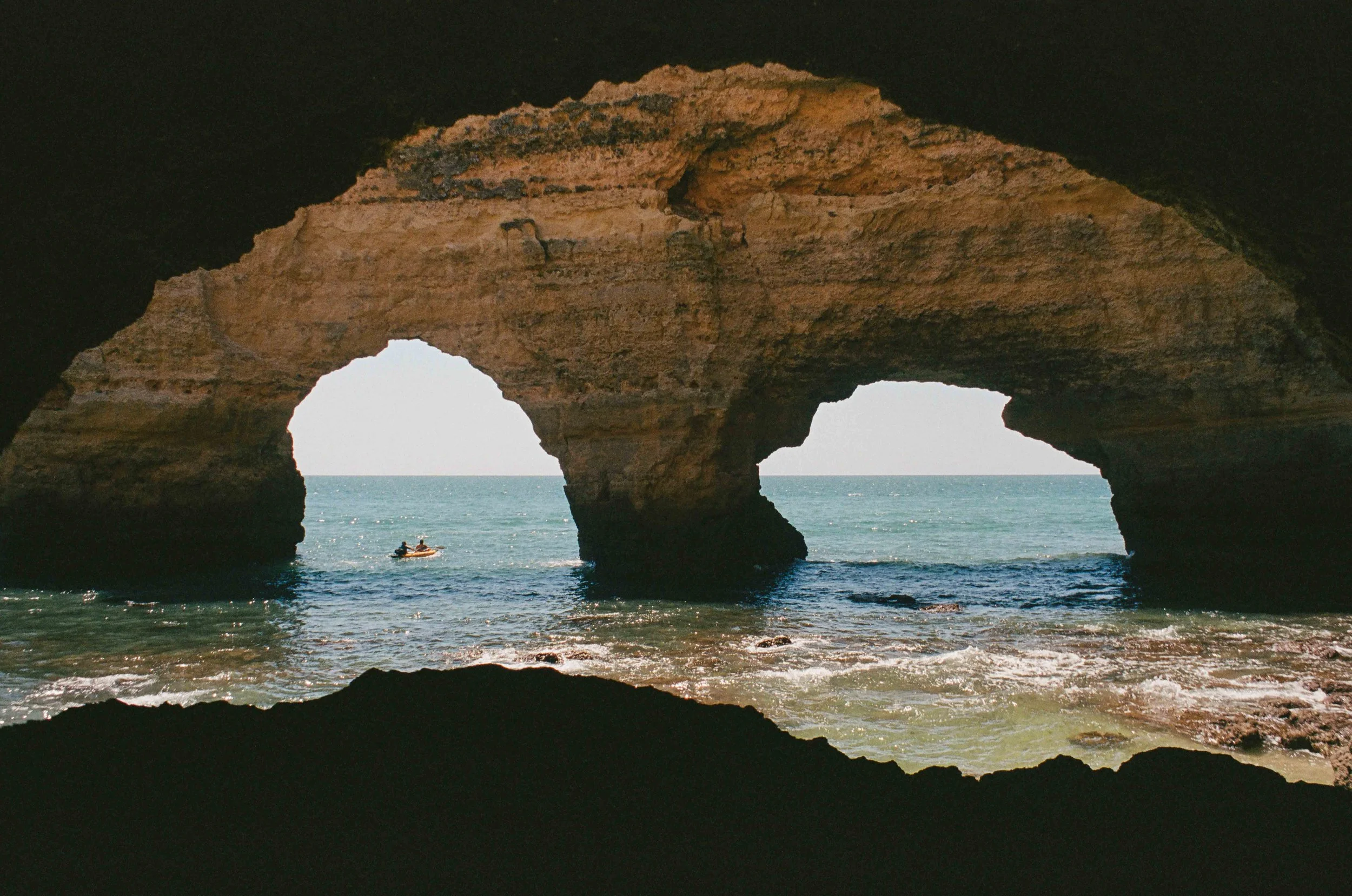 View of a beach with two natural rock arches seen from inside a cave, with water and a small boat in the background.