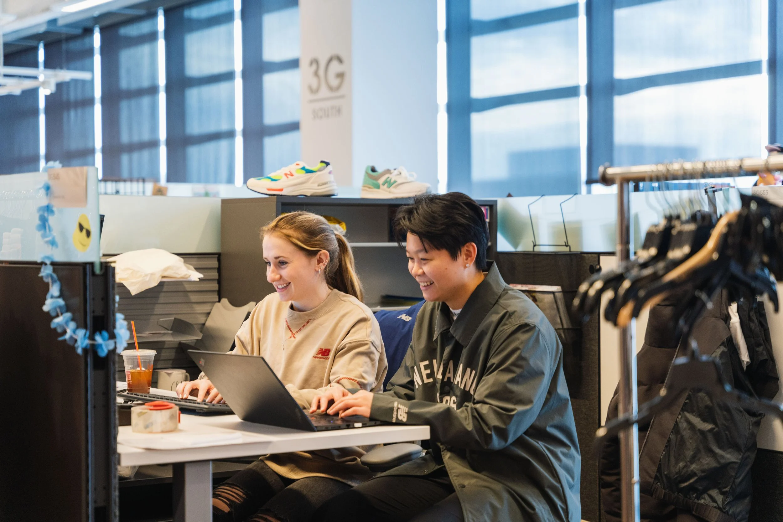 Two smiling young adults, a woman and a man, sitting at a desk in an office, working on laptops, with a windowed building in the background.