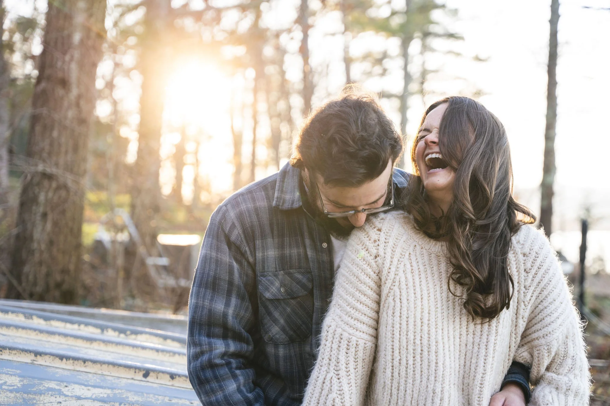 A couple sharing a laugh outdoors in a wooded area during sunset.