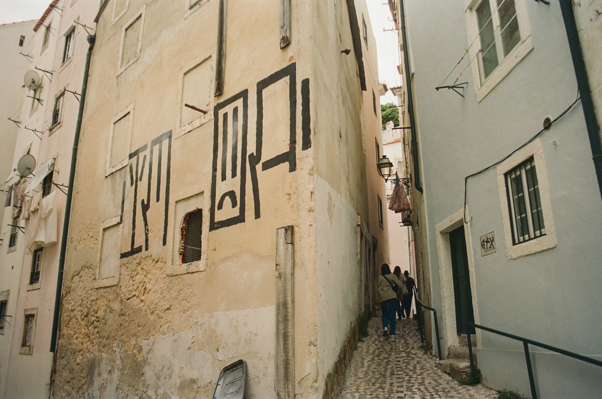 Narrow alleyway between old buildings with pastel-colored walls, laundry hanging on lines, and a small group of people walking.