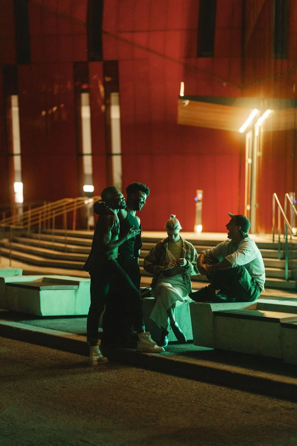 Four young adults socializing outdoors at night, sitting and standing on concrete benches, with a modern building with red walls and bright lights in the background.