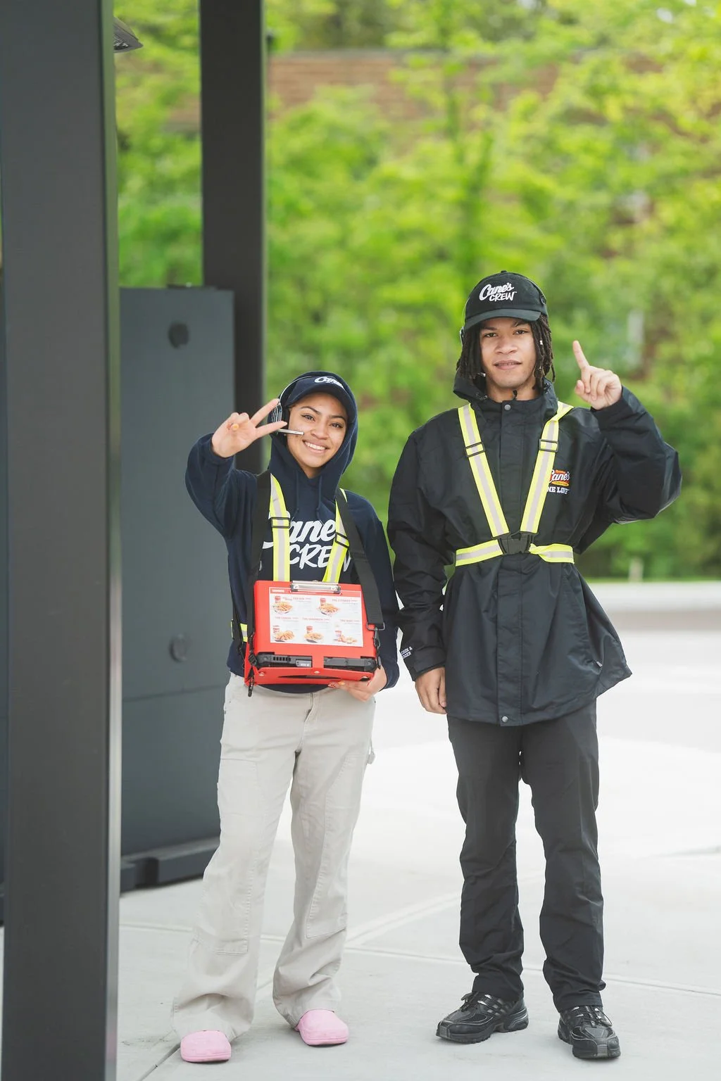 Two smiling food delivery workers standing outside, wearing black uniforms with yellow reflective straps, and black caps with a logo. One holds a menu and makes a peace sign, the other points upward, with a green, leafy background.